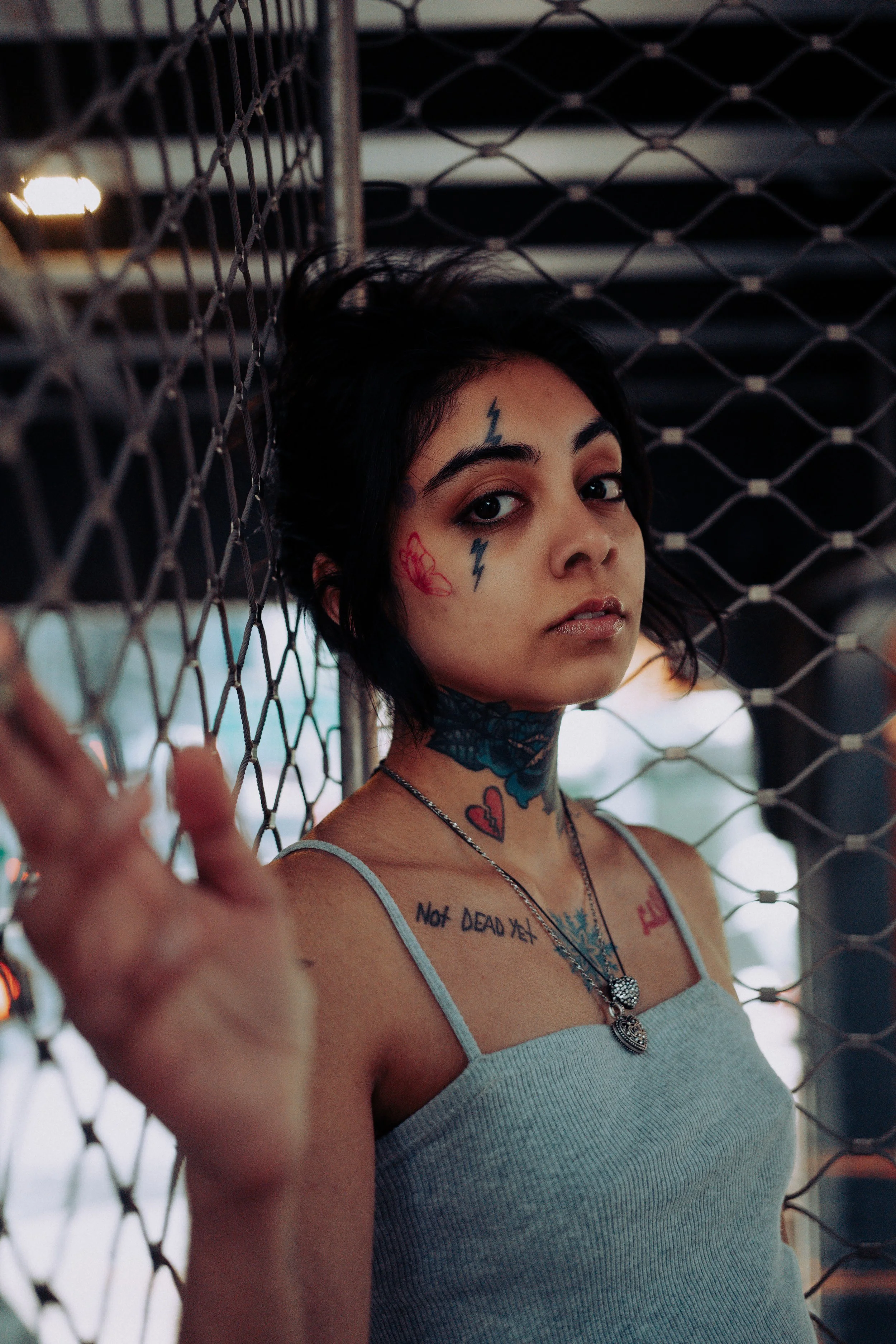 A young woman with tattoos on her face and neck. She has dark hair and is wearing a light-colored tank top and necklaces. She is standing behind a chain-link fence, looking directly at the camera with a serious expression.