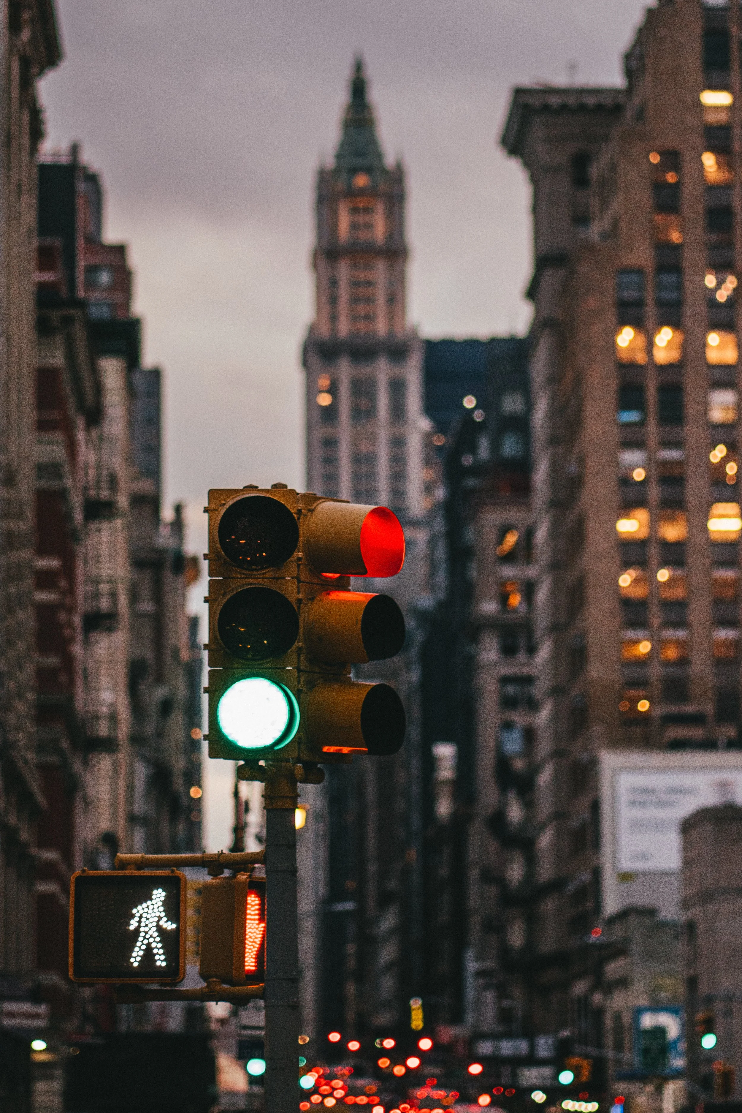 City street scene during dusk with a traffic light showing green and a pedestrian crossing light. Tall buildings with illuminated windows line the street and a blurred clock tower is visible in the background.