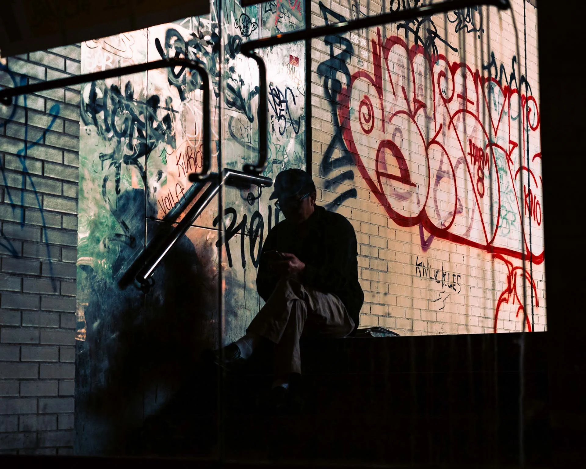 A man sits outside on his phone, beyond a glass door. The wall behind him is covered in graffiti. The light and shadow of the image give the viewer a unique perspective.