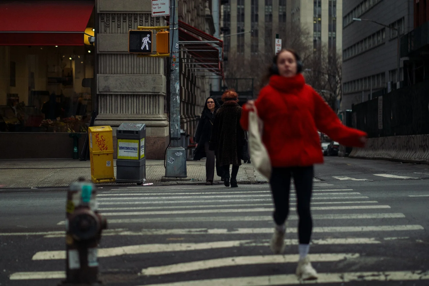 Blurred woman in a red jacket crossing as another woman watches from the corner