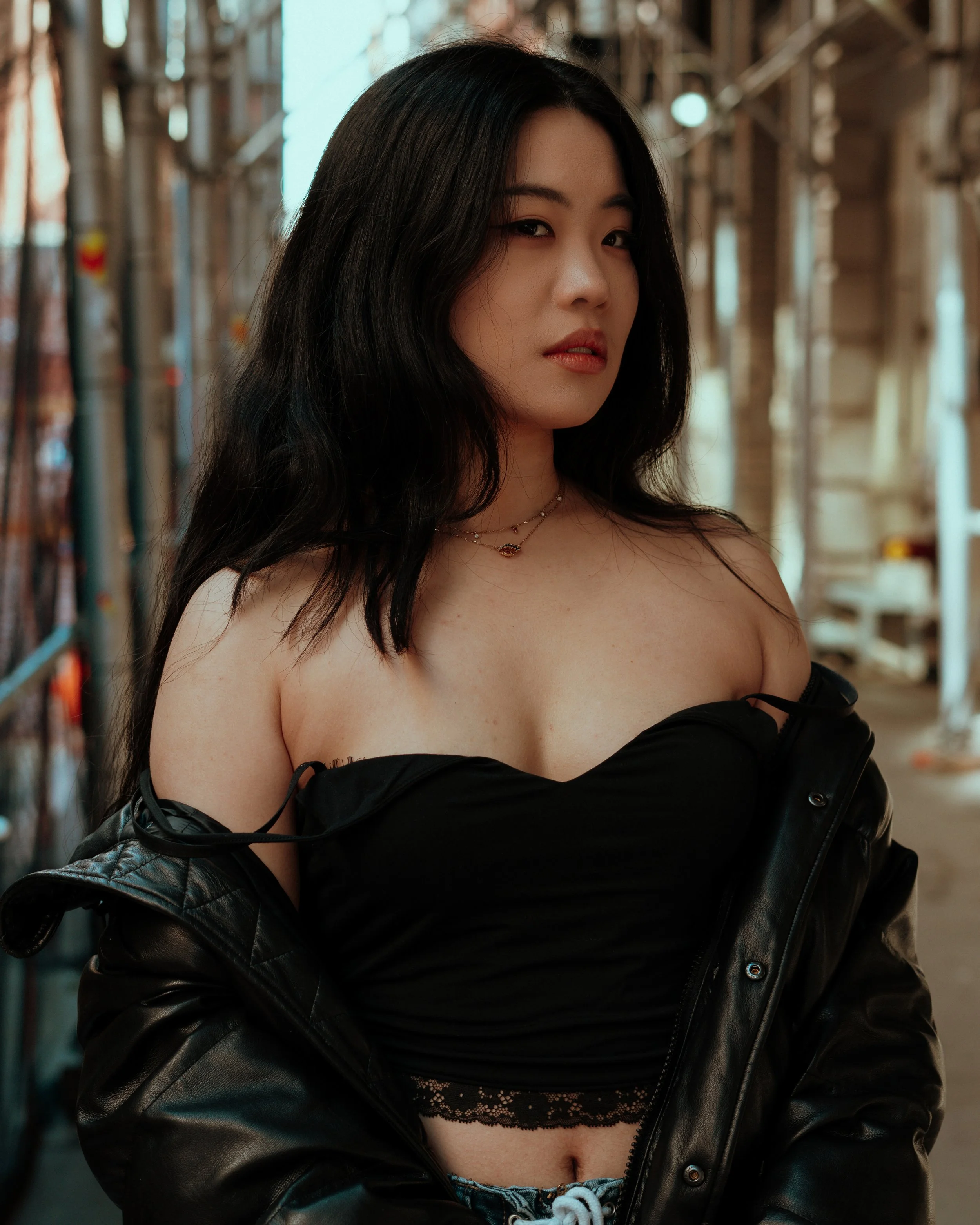 A young Asian woman with long black hair gazes at the camera, standing urban New York City street. She wears a black off-shoulder top, lace bralette, and a black leather jacket draped over her arms, with layered necklaces around her neck.