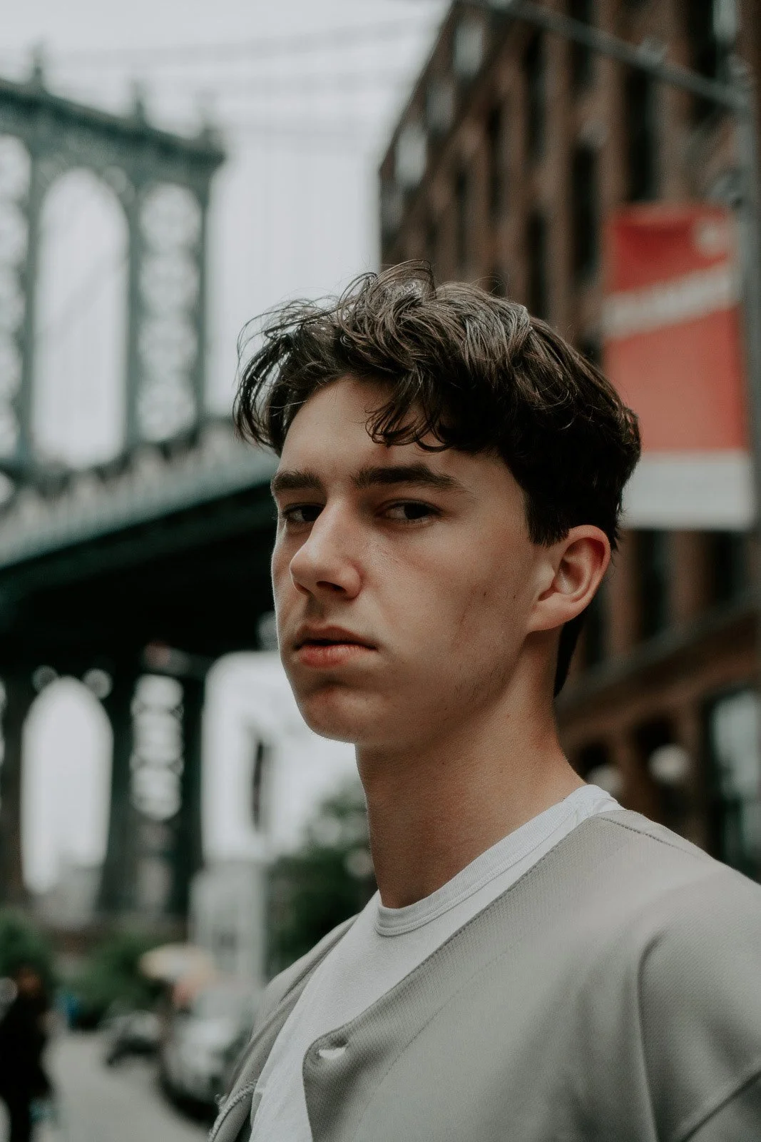 Close-up of a young man with dark wavy hair, standing outdoors in an urban environment with bridges and buildings in the background.