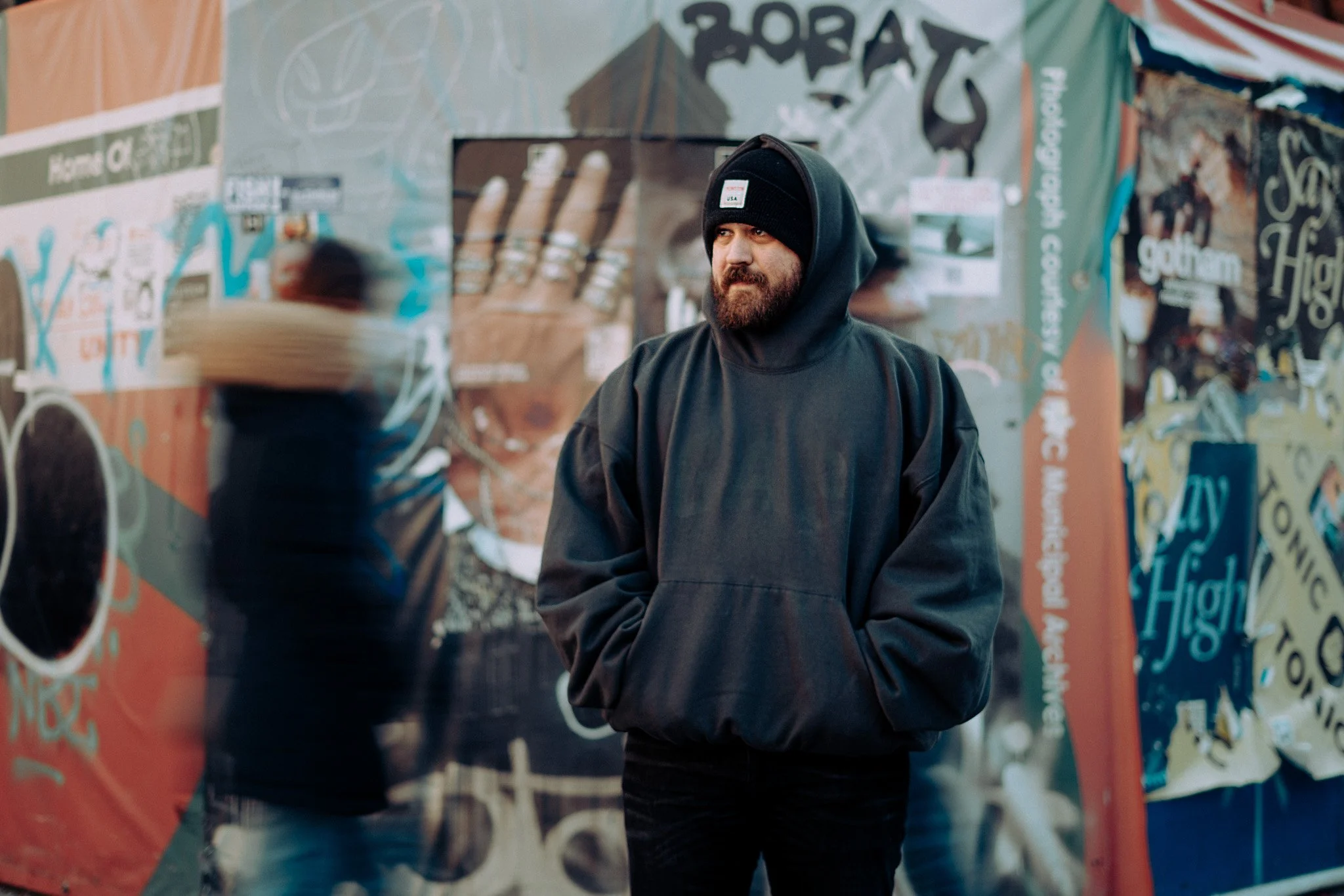 A man wearing a black hoodie and beanie stands in front of a colorful, graffiti-covered wall, looking to his right with a serious expression.
