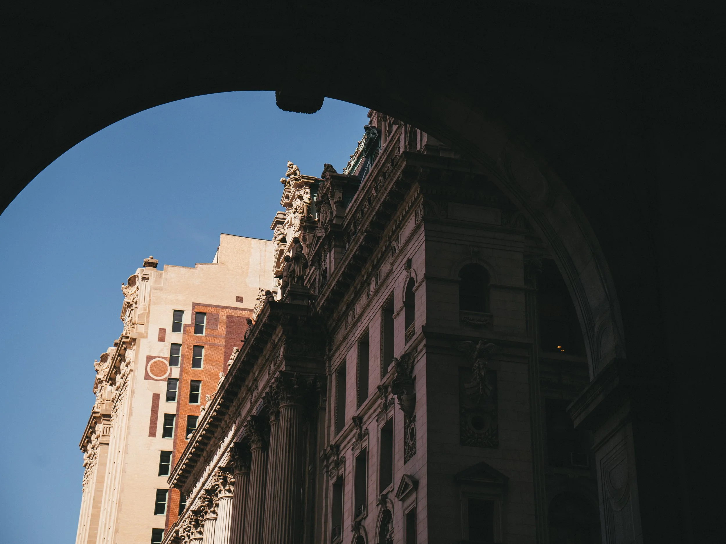 Looking up at ornate historic buildings with classical architecture, including decorative sculptures and columns, through a dark archway against a clear blue sky.