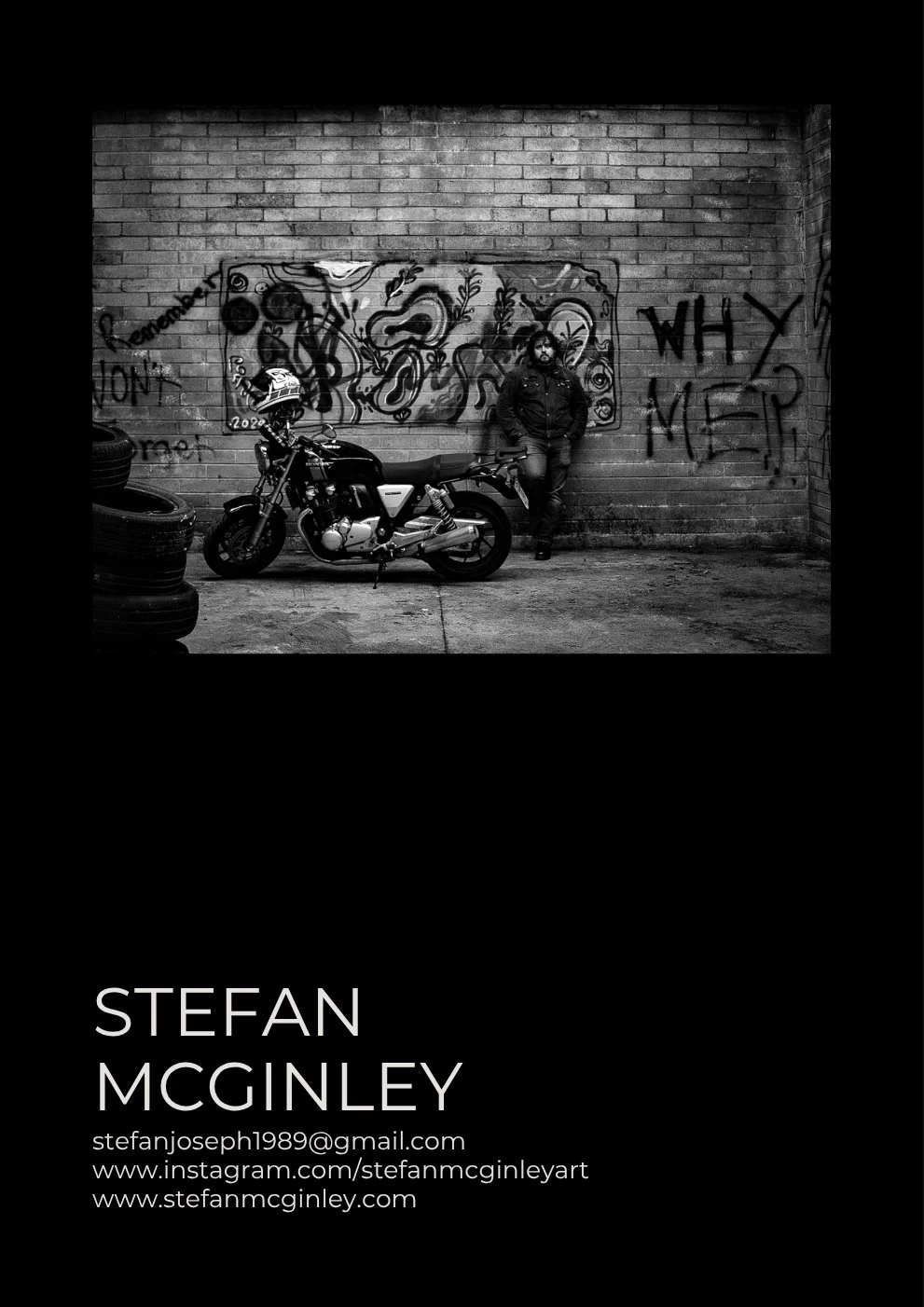 Black and white photograph of a man with long hair and beard leaning against a graffiti-covered brick wall, next to a motorcycle with a helmet hanging on its handlebar, and stacked tires in the foreground.