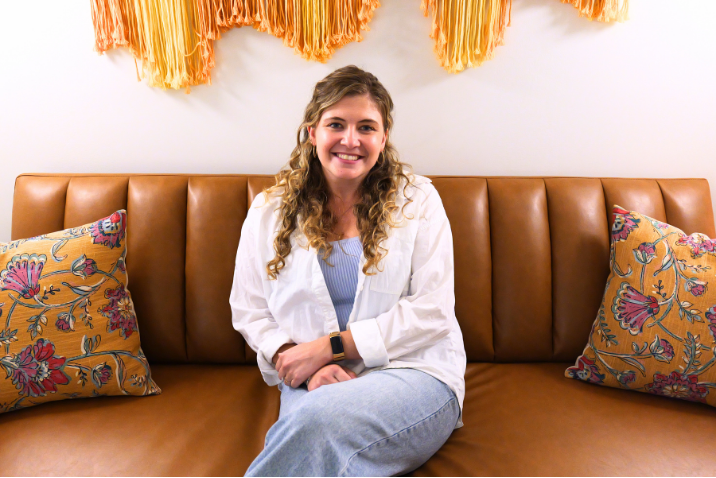 A young woman with curly hair sitting on a tan leather couch, smiling, with colorful pillows and a textured orange wall hanging behind her.