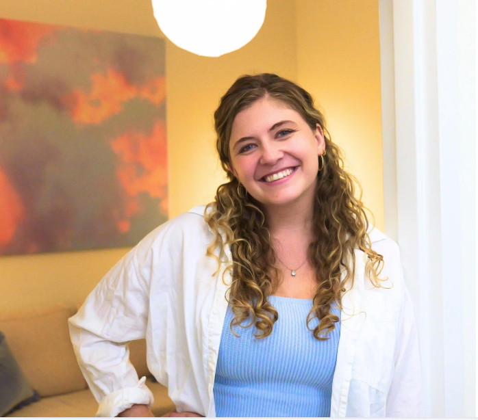 A young woman with long curly hair is smiling at the camera, wearing a white shirt over a light blue top, standing in a well-lit room with yellow walls, an abstract painting, and a round ceiling light.