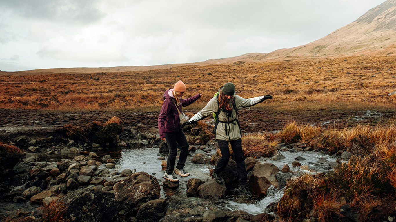 Two women hike over a rocky stream in a vast, open, grassy landscape with hills in the background.