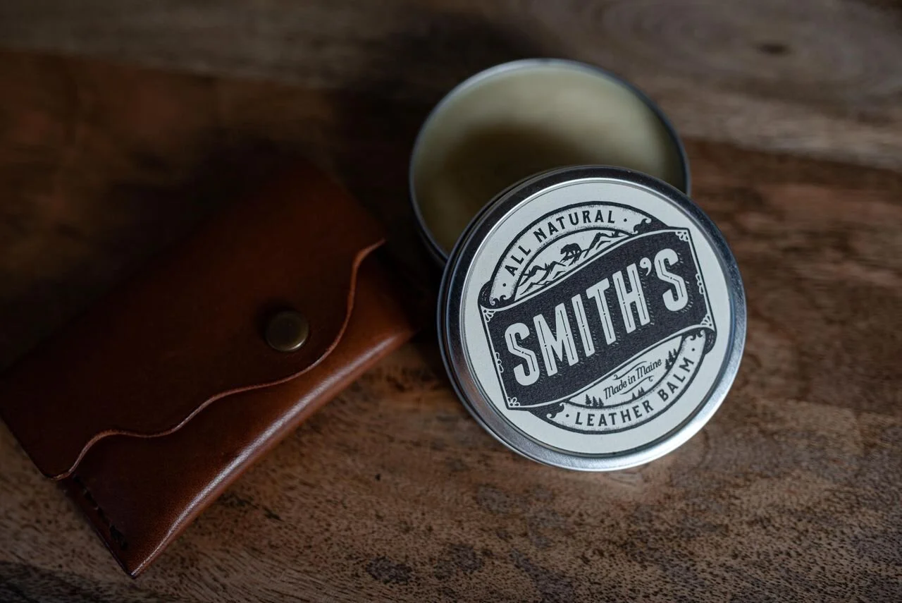 Tin of Smith's all natural leather balm on wooden surface with brown leather pouch.