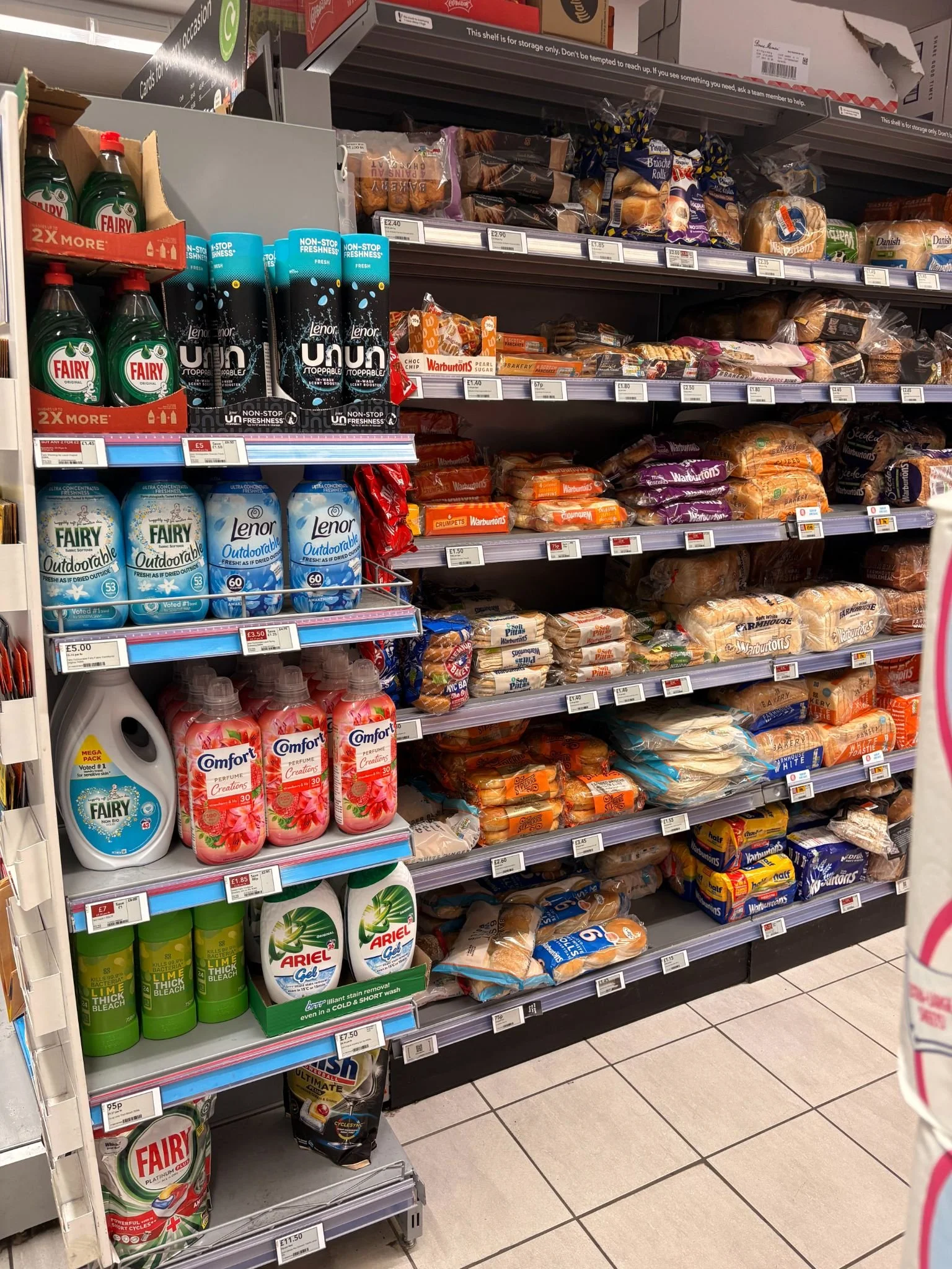 Supermarket shelf with various bread and bakery products, including loaves of bread, packaged buns, and bread rolls.