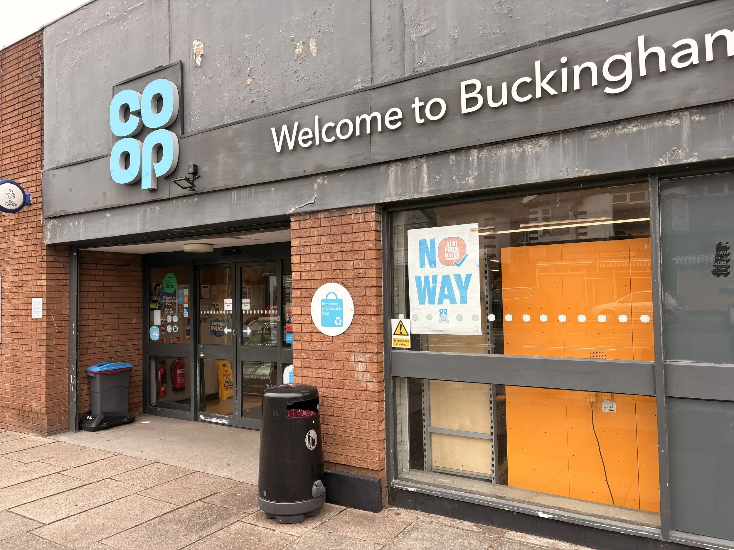 The exterior of a Coop grocery store with a sign that reads "Welcome to Buckingham". There is an entrance door with various stickers, a trash bin, and an orange-colored interior visible through the window. A sign near the door reminds customers to remember reusable bags.