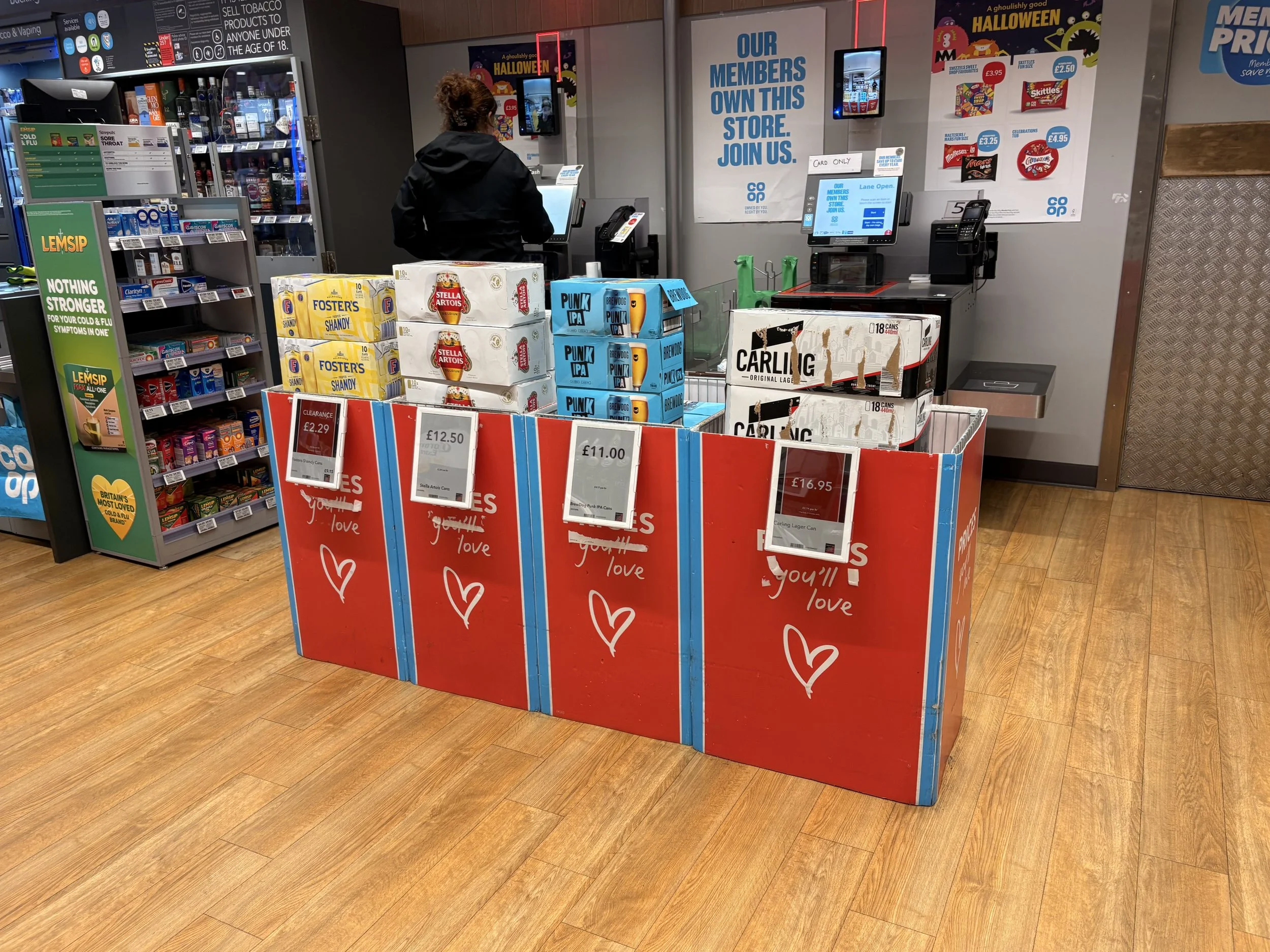 Display of various canned alcoholic beverages in a store, with price tags and a cashier in the background.