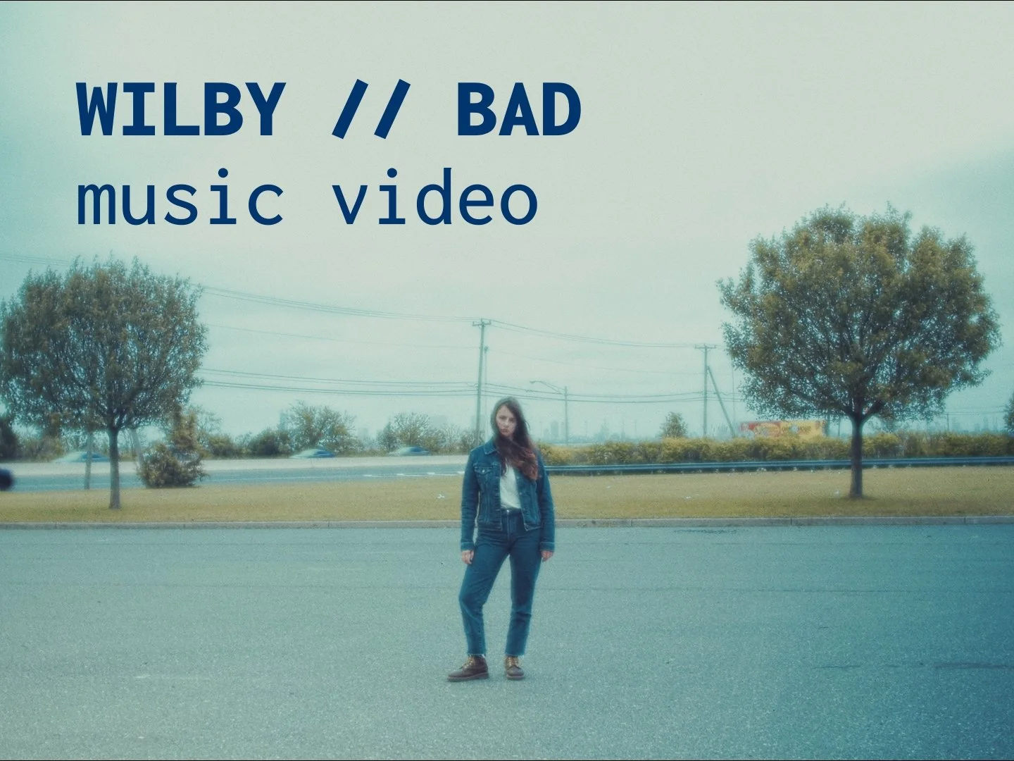 A woman standing in an empty parking lot under a cloudy sky, with two trees, power lines, and a road in the background, overlaid with text 'WILBY // BAD music video'.