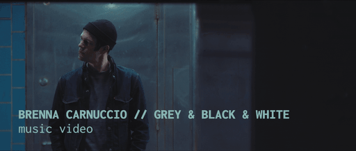 A man wearing a dark jacket and beanie standing against a grey background with blue lighting, with text overlay indicating Brenna Carnuccio, Grey & Black & White, music video.