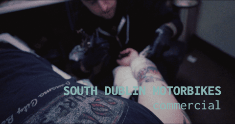A tattoo artist is drawing on a person's leg in a tattoo studio, with the words 'South Dublin Motorbikes' visible in the background.