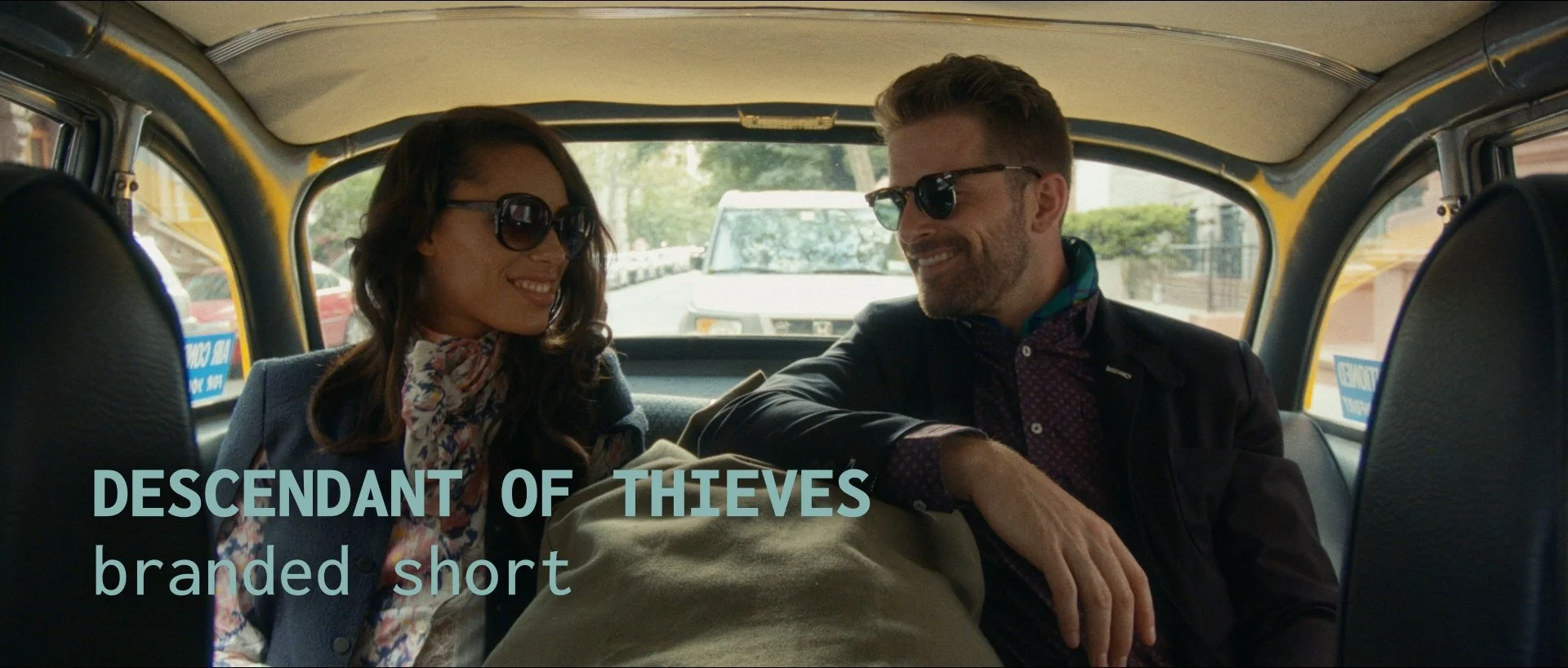 A man and woman sitting in the back of a taxi, smiling at each other. They are both wearing sunglasses and casual clothes.