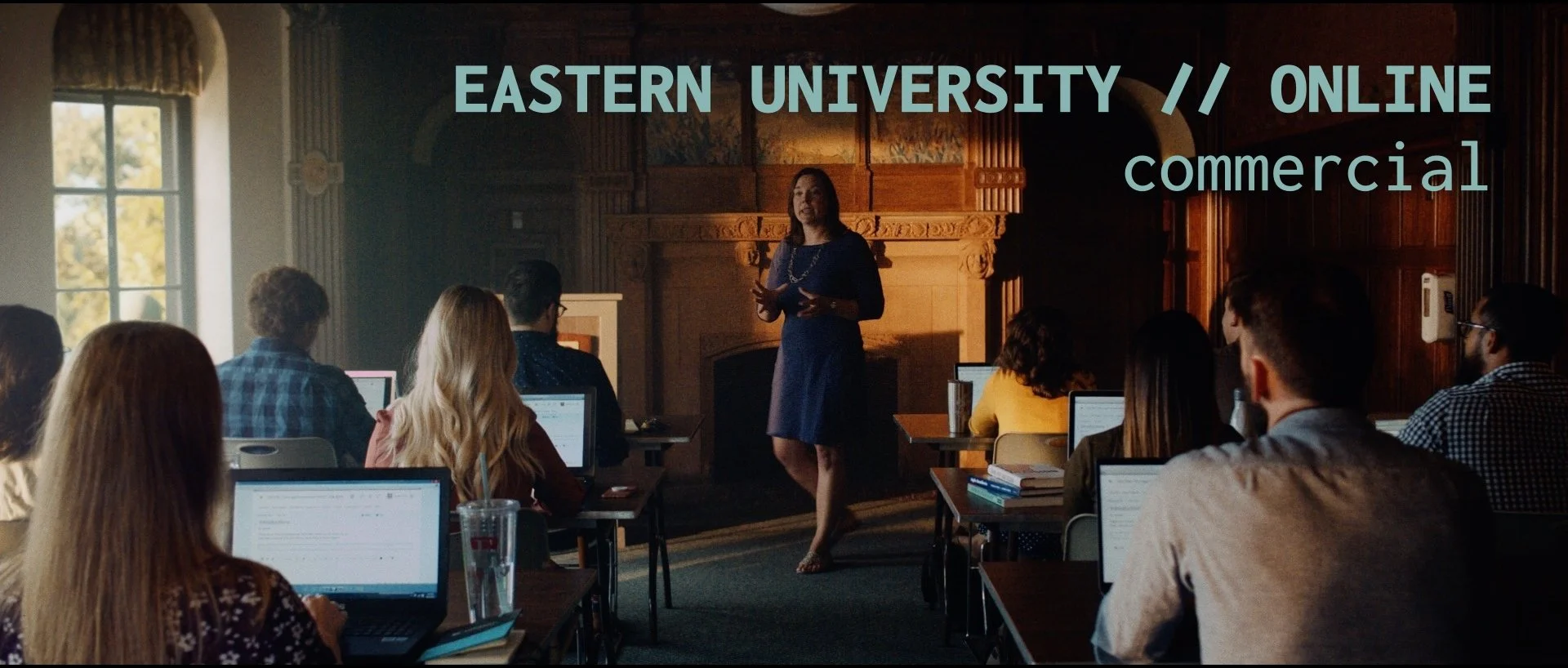 A woman standing and speaking to a classroom of students with laptops open, in a room with wood-paneled walls and large windows, during an online commercial class at Eastern University.