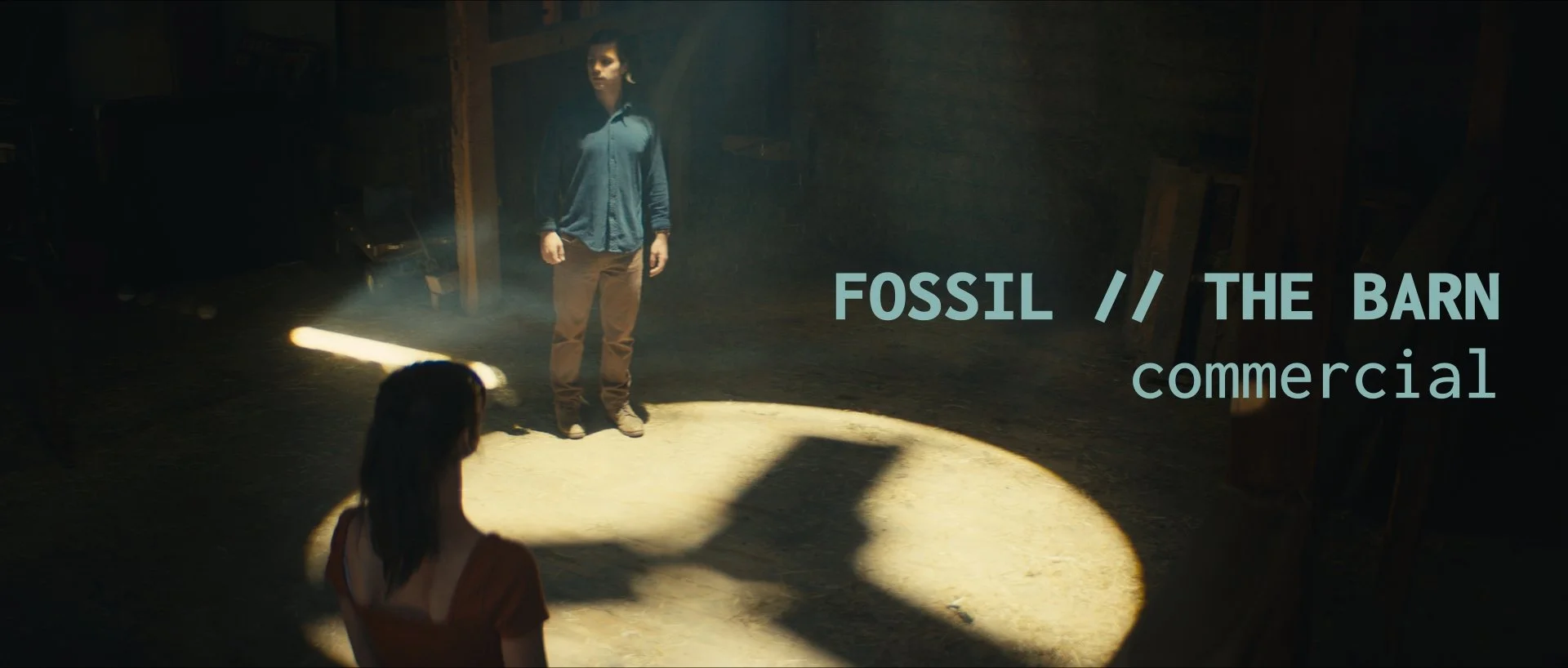 A dimly lit barn interior with a woman in red facing a woman in a blue shirt and brown pants standing in a spotlight. Text overlay reads 'FOSSIL // THE BARN commercial'.