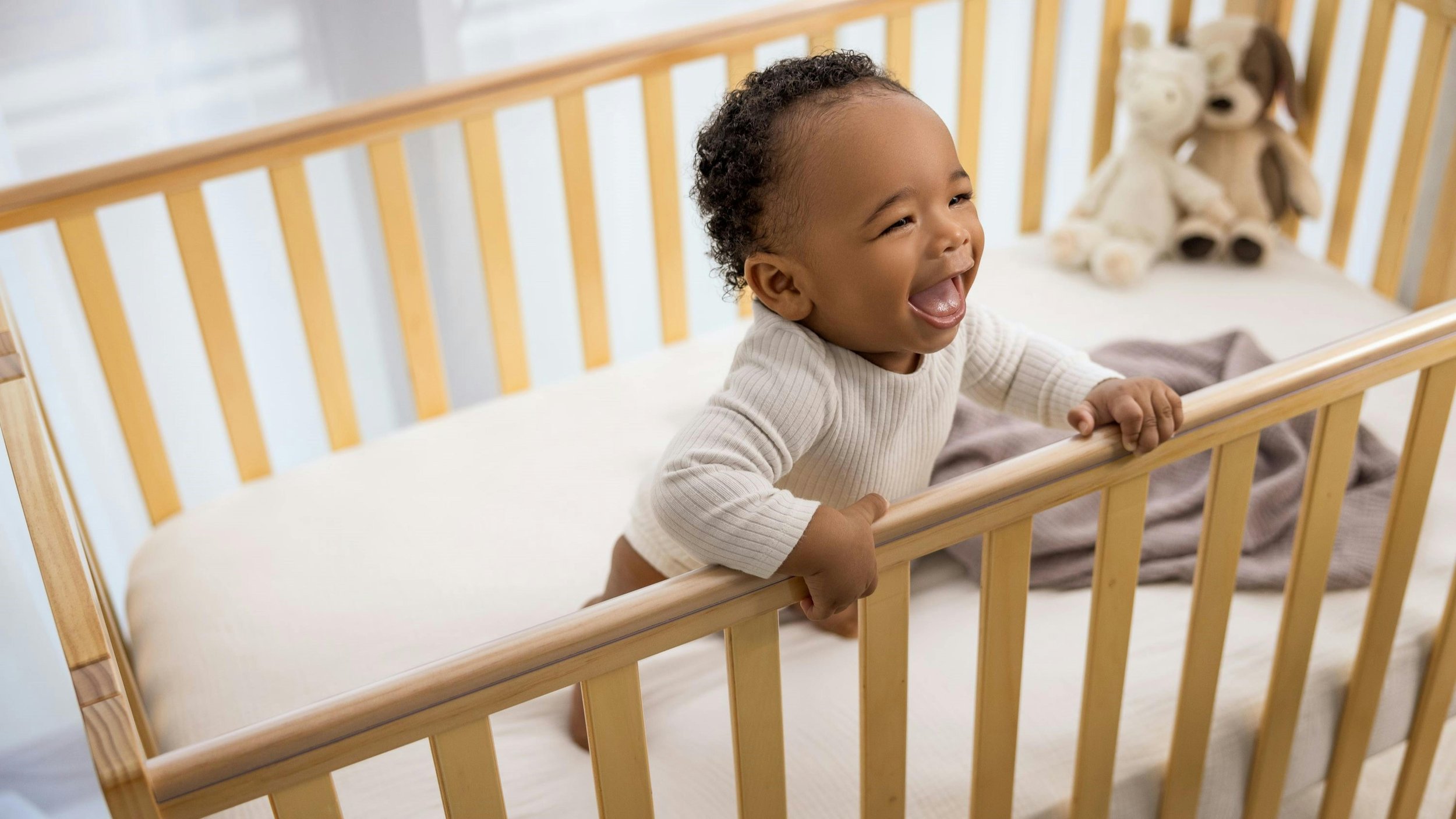 Baby standing in their cot, holding the rails, looking happy and alert