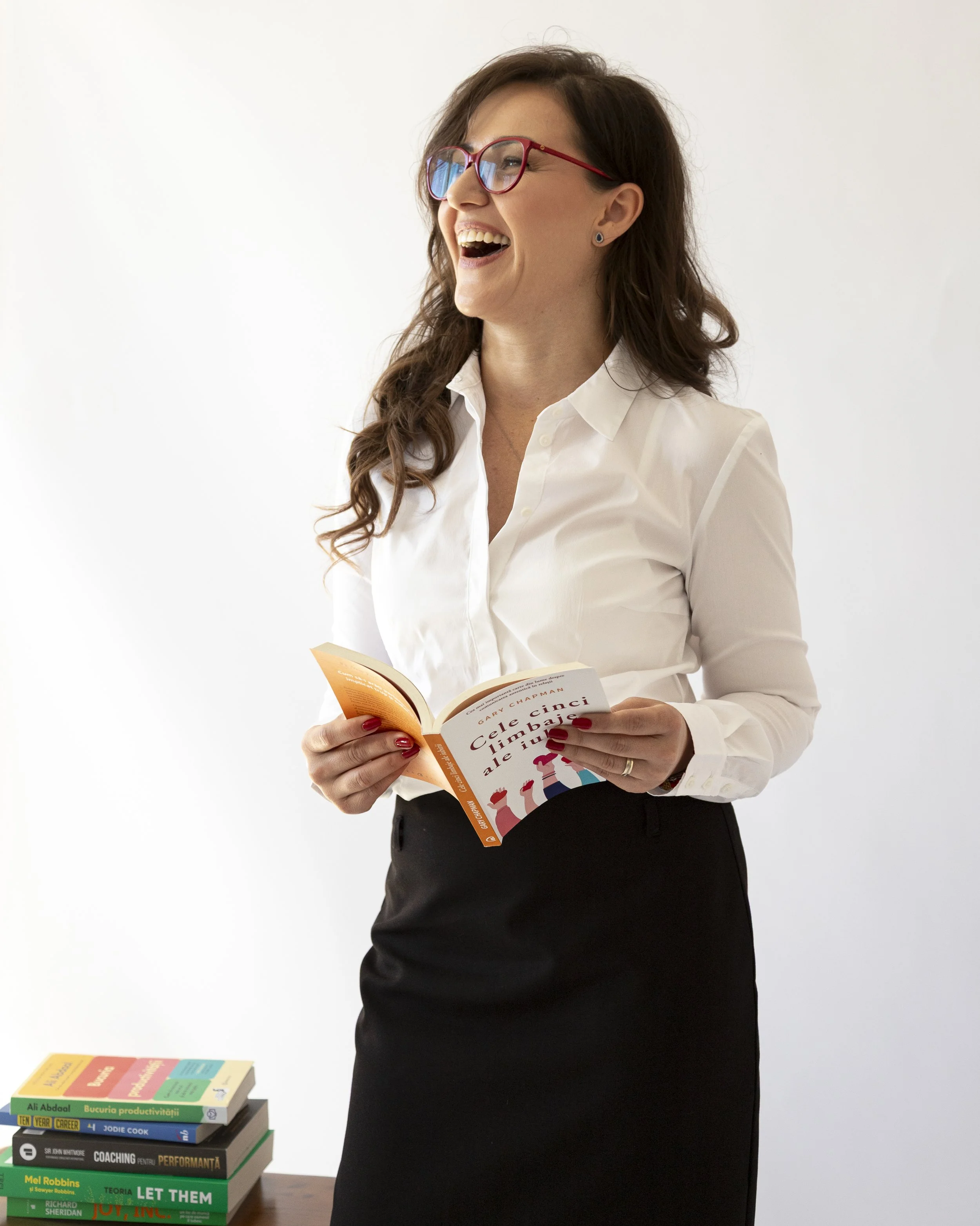 Smiling woman with glasses reading a book, standing next to a stack of books on a table.