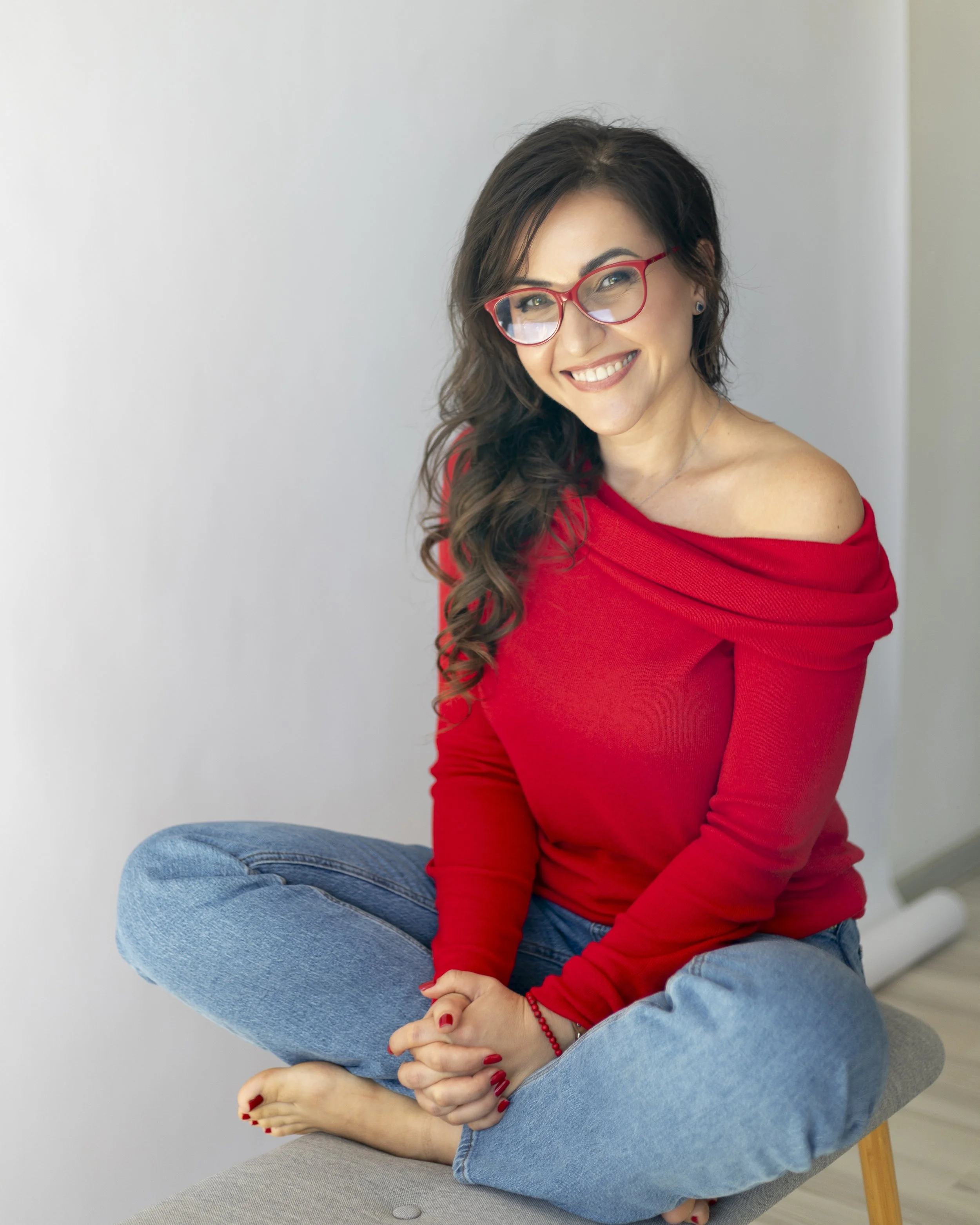 Woman with long dark wavy hair wearing red glasses, a red off-the-shoulder sweater, and blue jeans, sitting cross-legged on a gray bench, smiling at the camera.