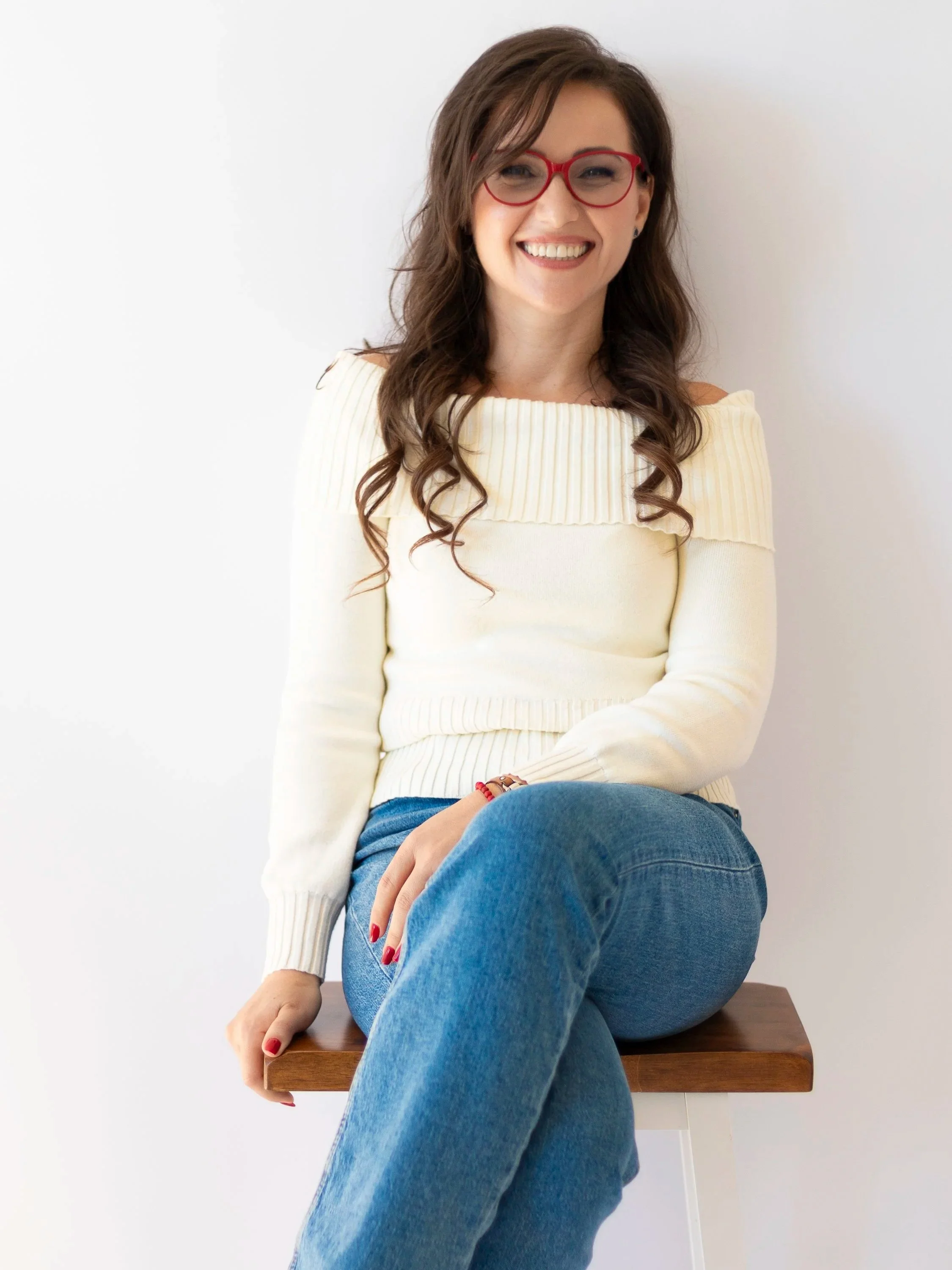 A woman with brown wavy hair, red glasses, and a warm smile, wearing a cream-colored off-the-shoulder sweater and blue jeans, sitting on a wooden stool against a plain white wall.