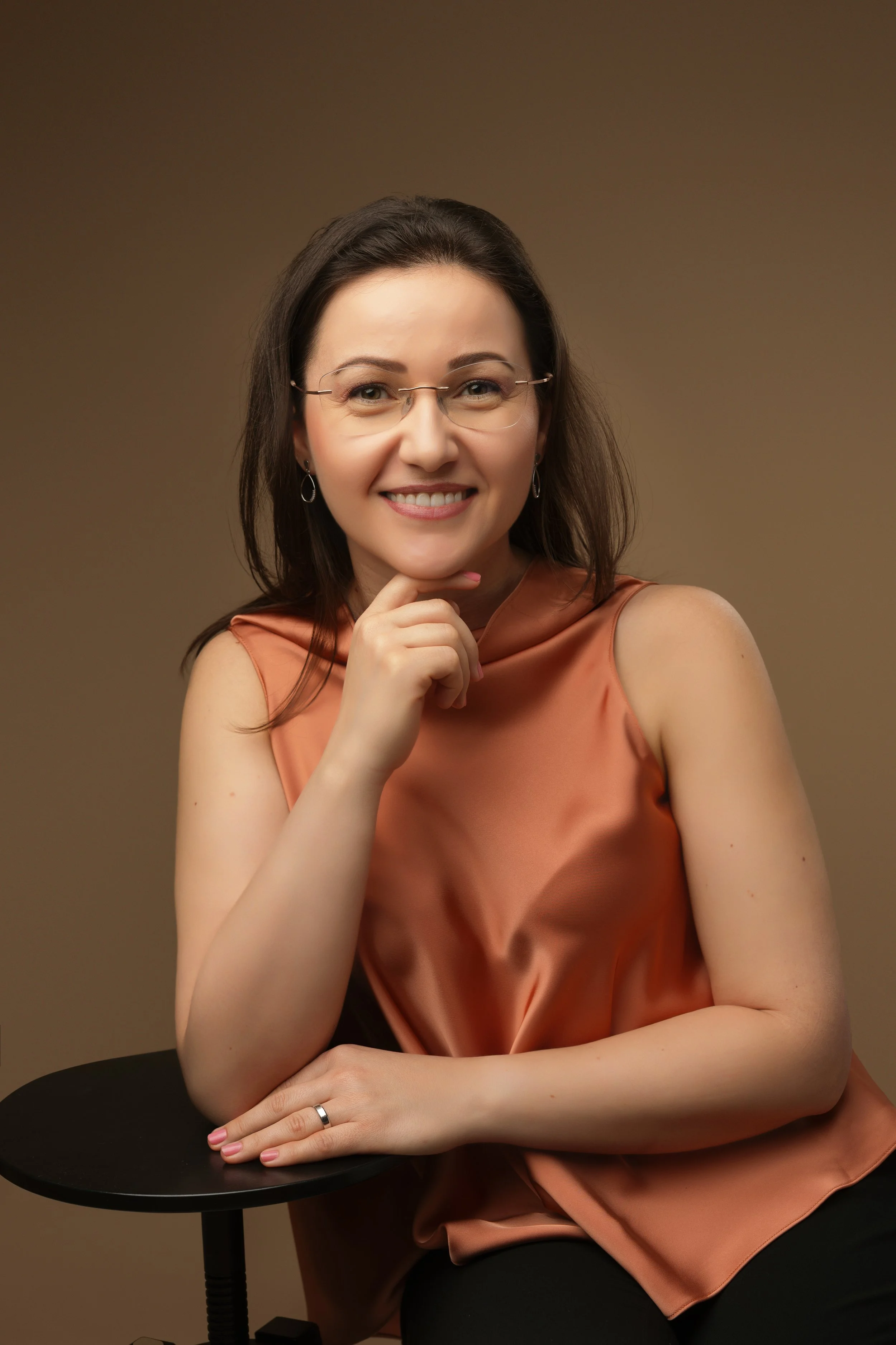A woman with brown hair, glasses, and earrings smiling and sitting with her chin on her hand against a plain gradient background.