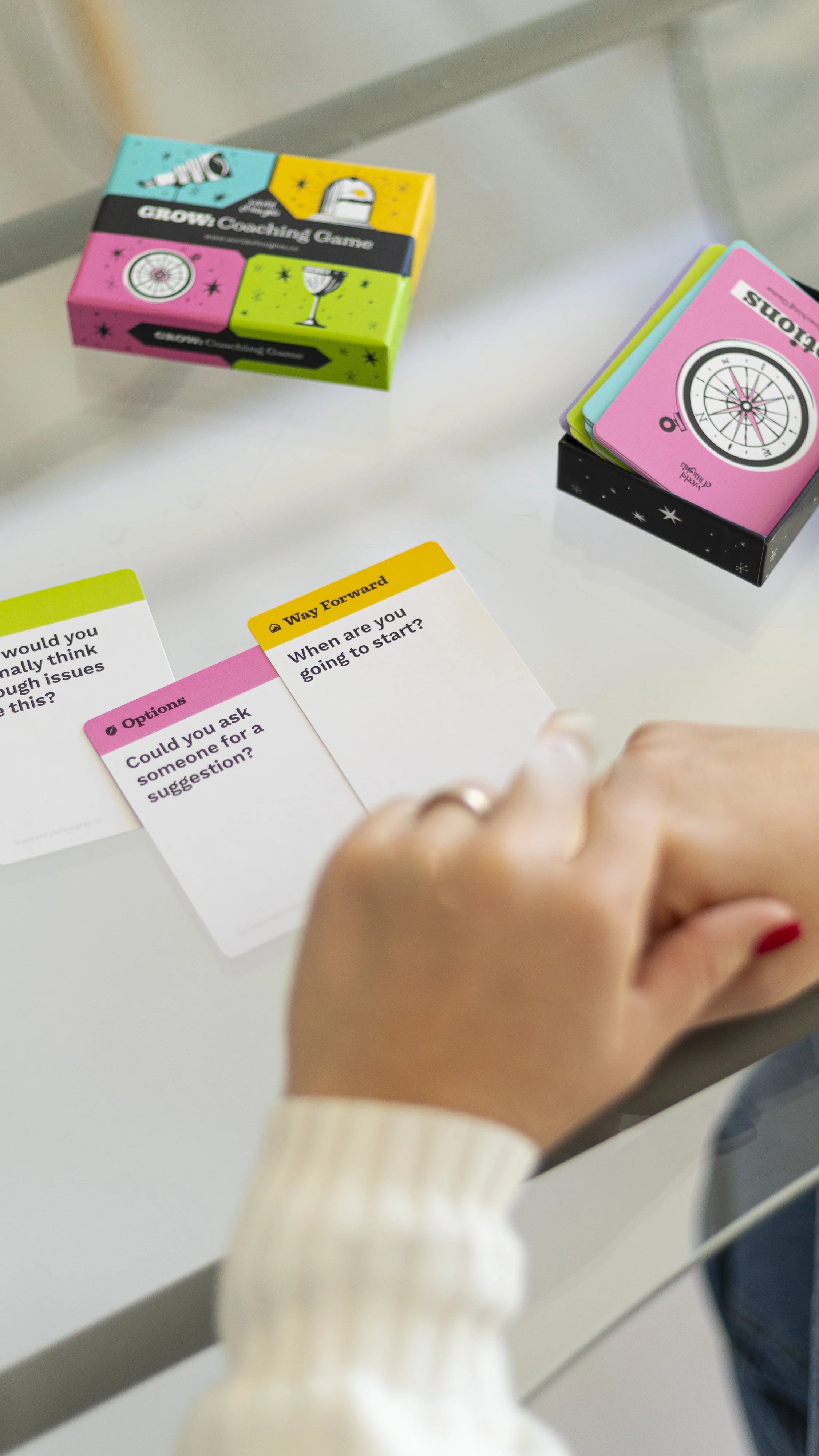 A person holding a card from a coaching game on a desk. There are colorful game boxes and cards with questions on the desk.