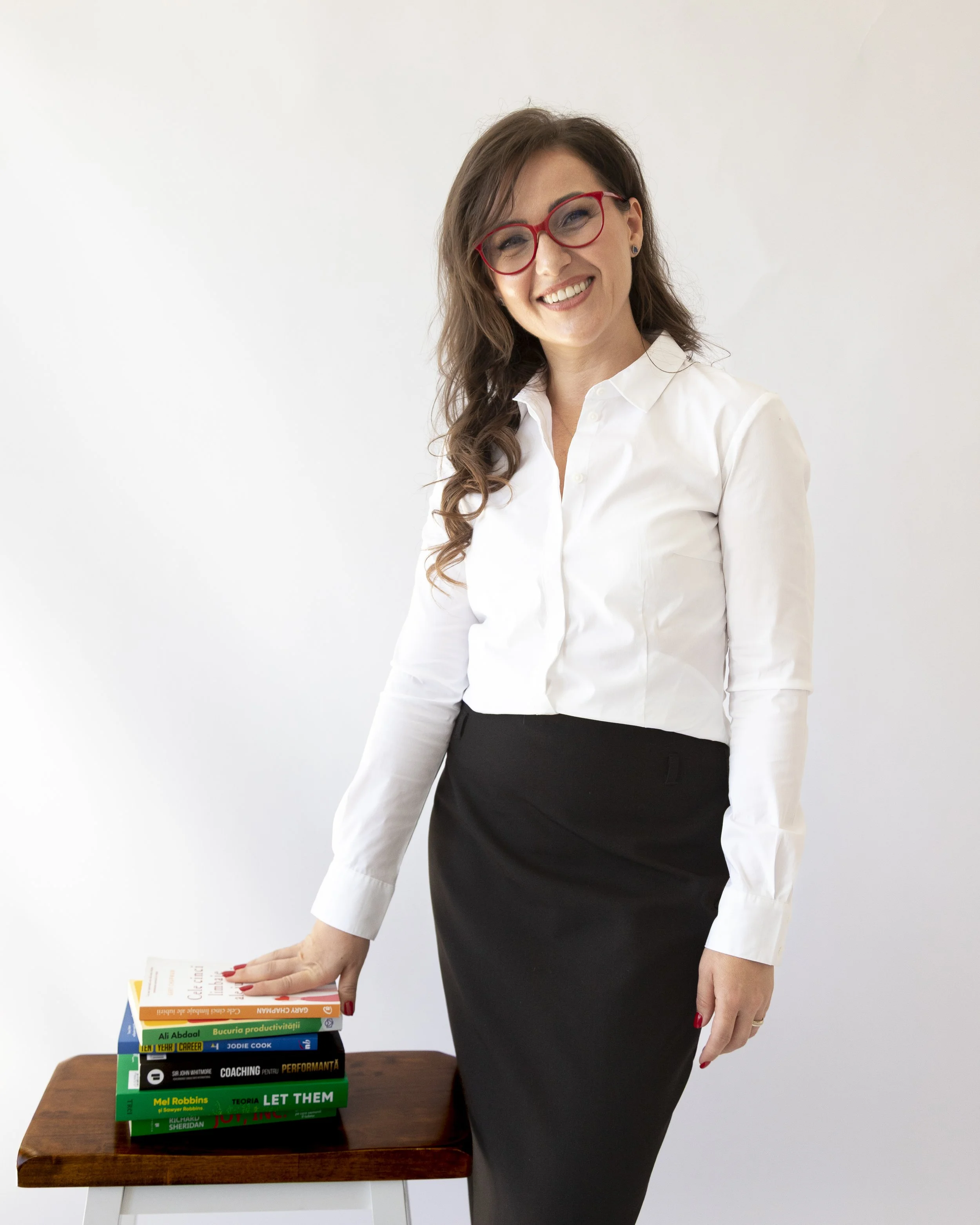A woman with long, wavy brown hair, wearing red glasses, a white blouse, and a black skirt, standing next to a wooden table with a stack of colorful books on it, smiling at the camera.