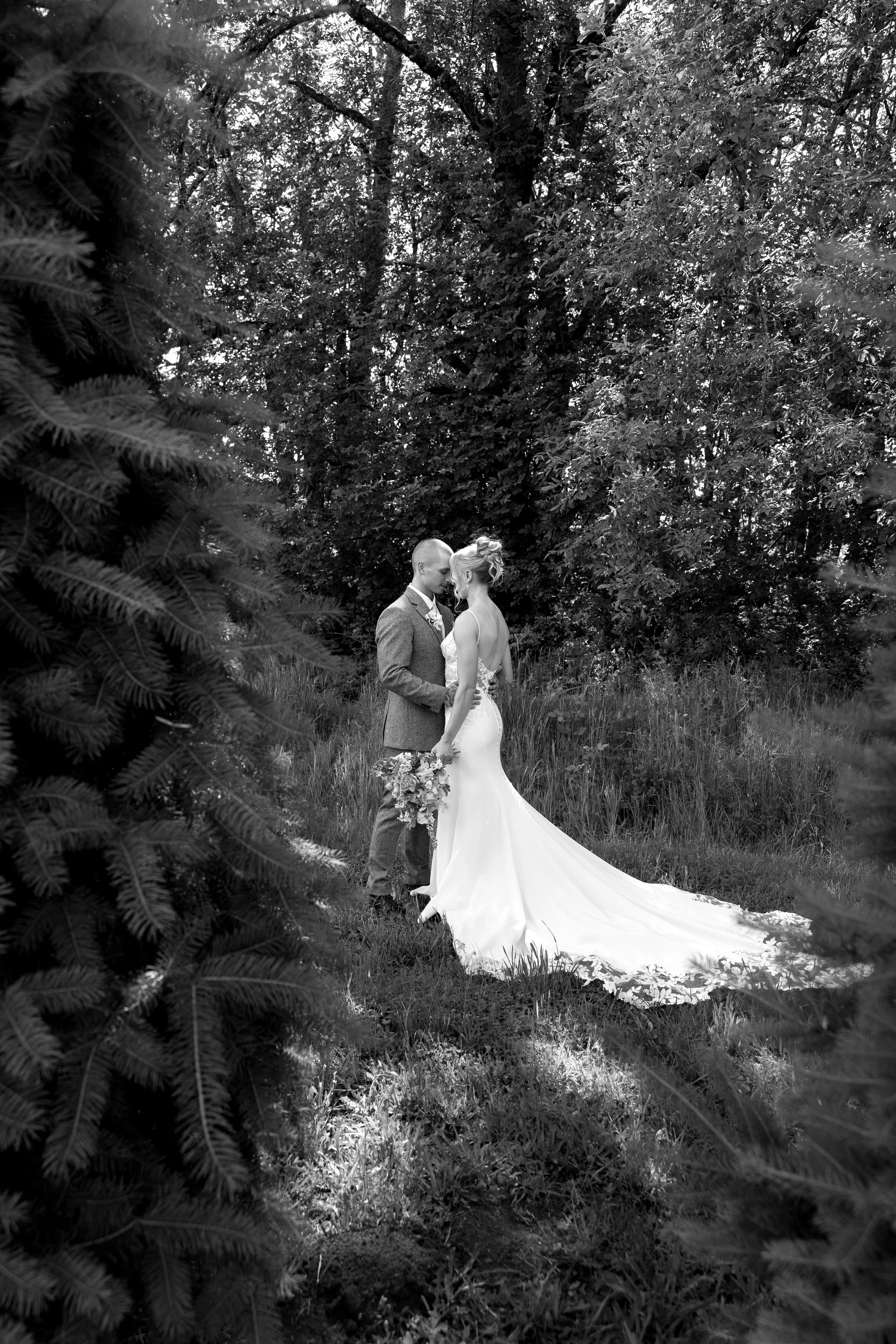 A bride and groom standing close together in a wooded outdoor setting, with the bride in a flowing wedding dress holding a bouquet, and the groom in a suit, touching foreheads.