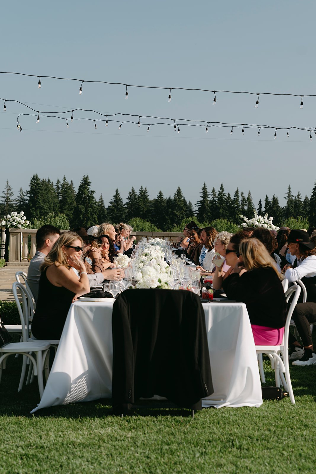 Outdoor wedding reception with a long white table, floral centerpieces, and guests seated on both sides, under string lights with trees in the background.