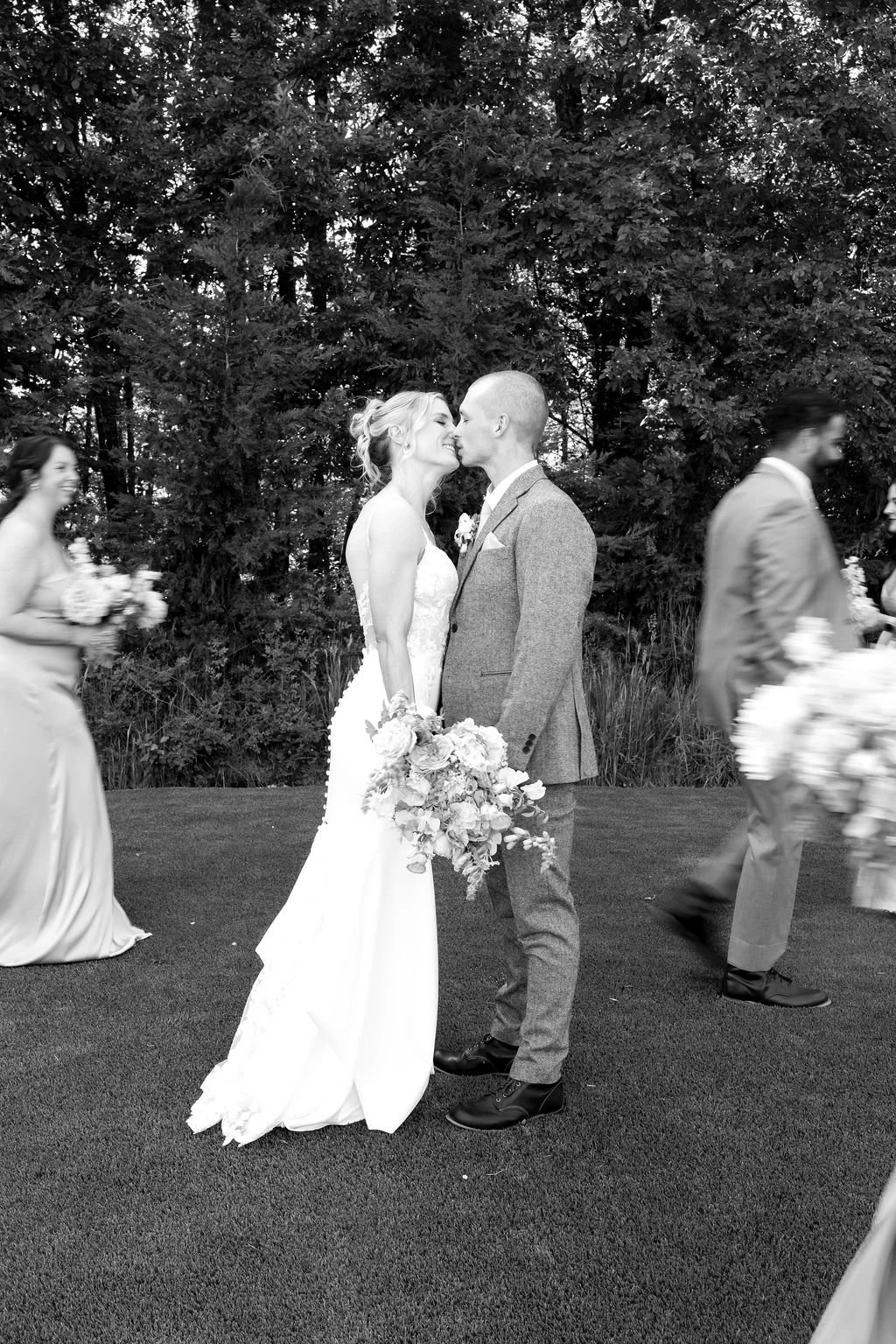 Black and white photo of a bride and groom kissing at their outdoor wedding ceremony, surrounded by friends holding bouquets and walking on a grassy area with trees in the background.