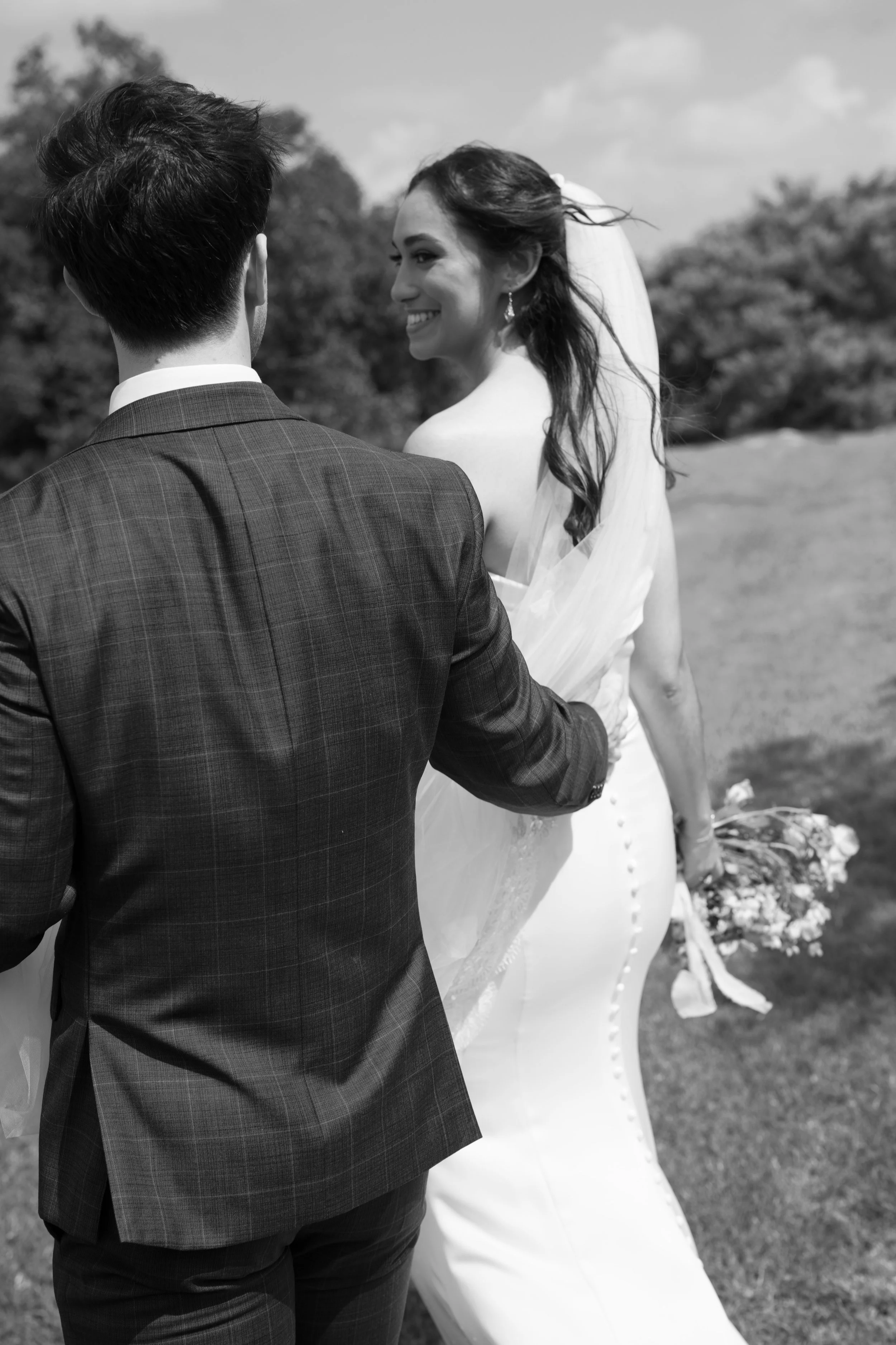 A black and white photo of a bride and groom outdoors, with the bride smiling and holding a bouquet of flowers, while the groom faces away.