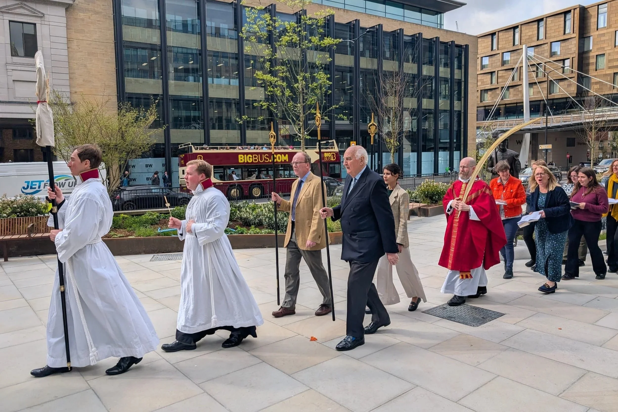 A religious procession with clergy and parishioners walking in a line on a city sidewalk, led by individuals dressed in white robes and carrying religious staff, followed by a man in a beige blazer and others in formal attire, with a priest in red ro