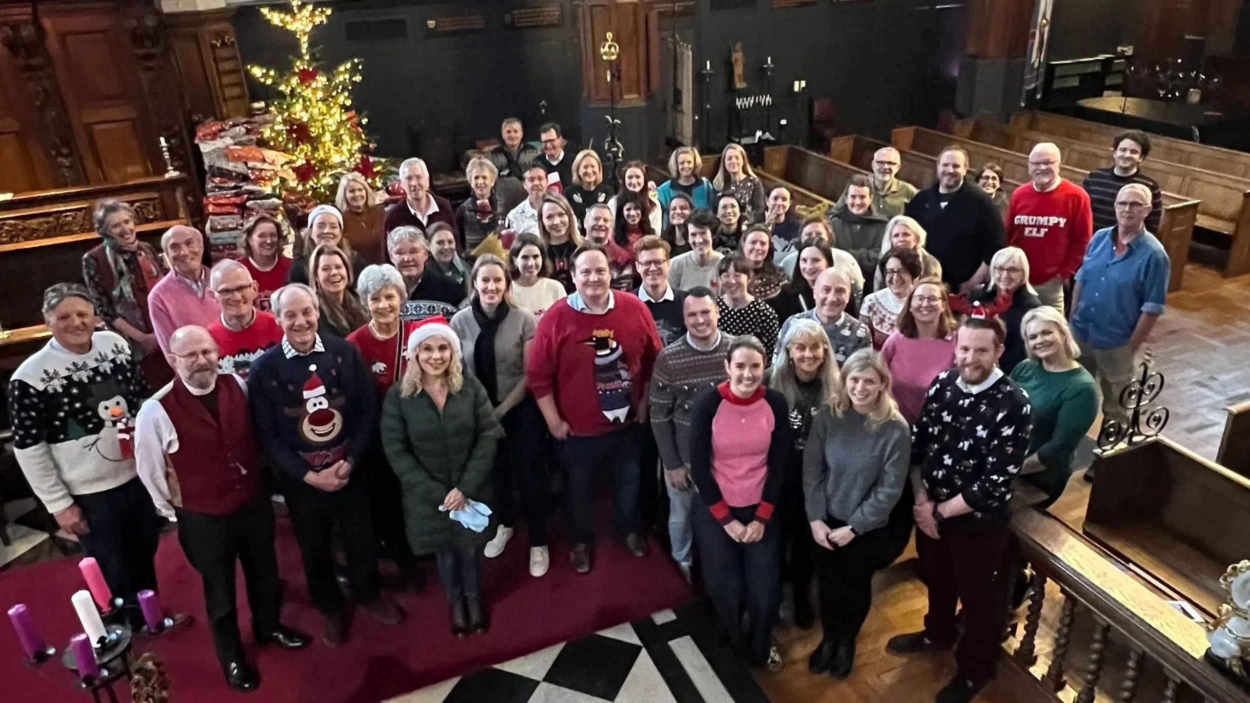 A large group of people gathered in a church decorated for Christmas, with a Christmas tree and wrapped presents in the background, all smiling at the camera.
