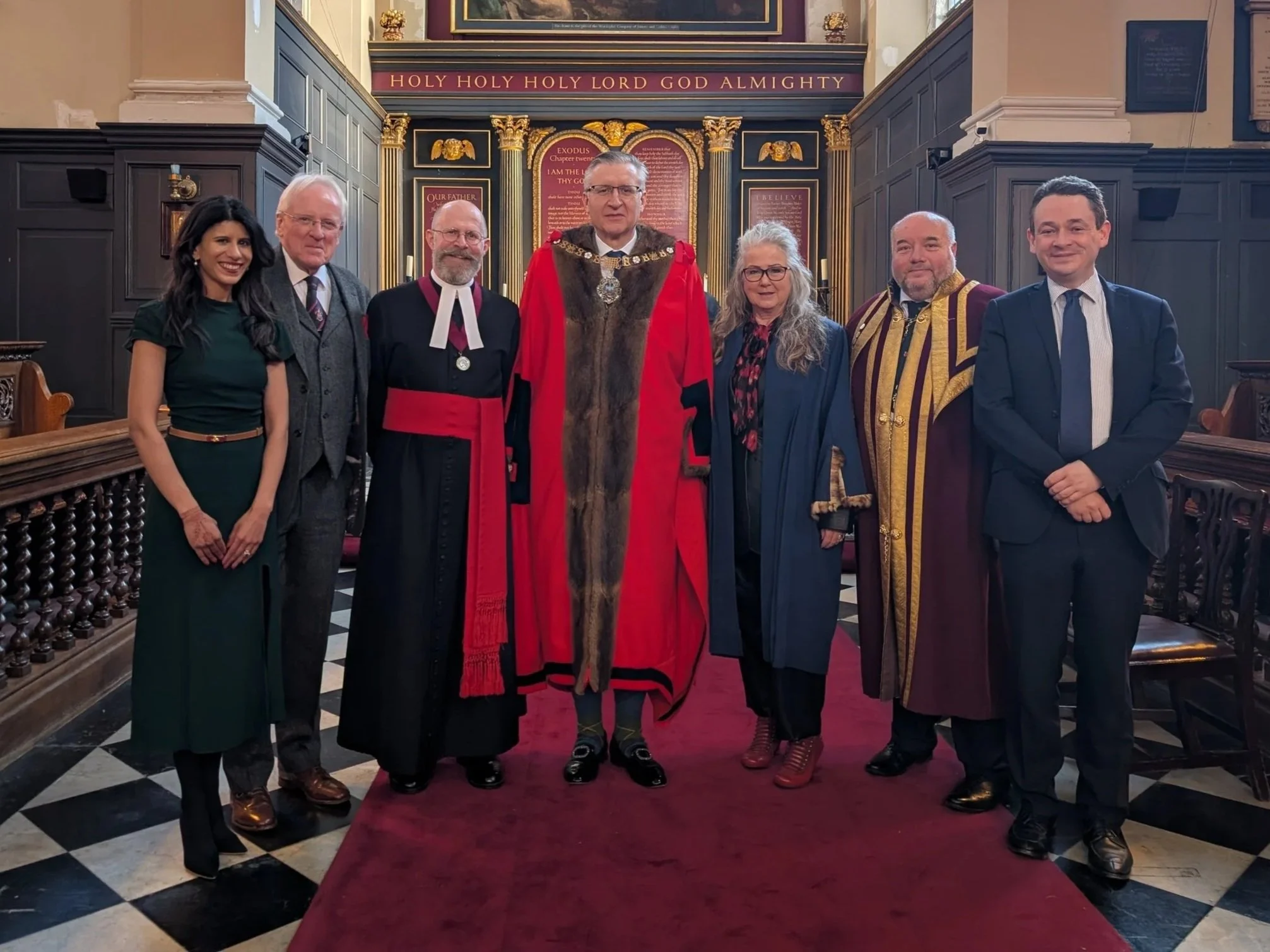 Group of seven people standing in a church, dressed in formal and academic robes, in front of an altar with a religious painting and inscriptions, inside a historic church with large stained glass windows.