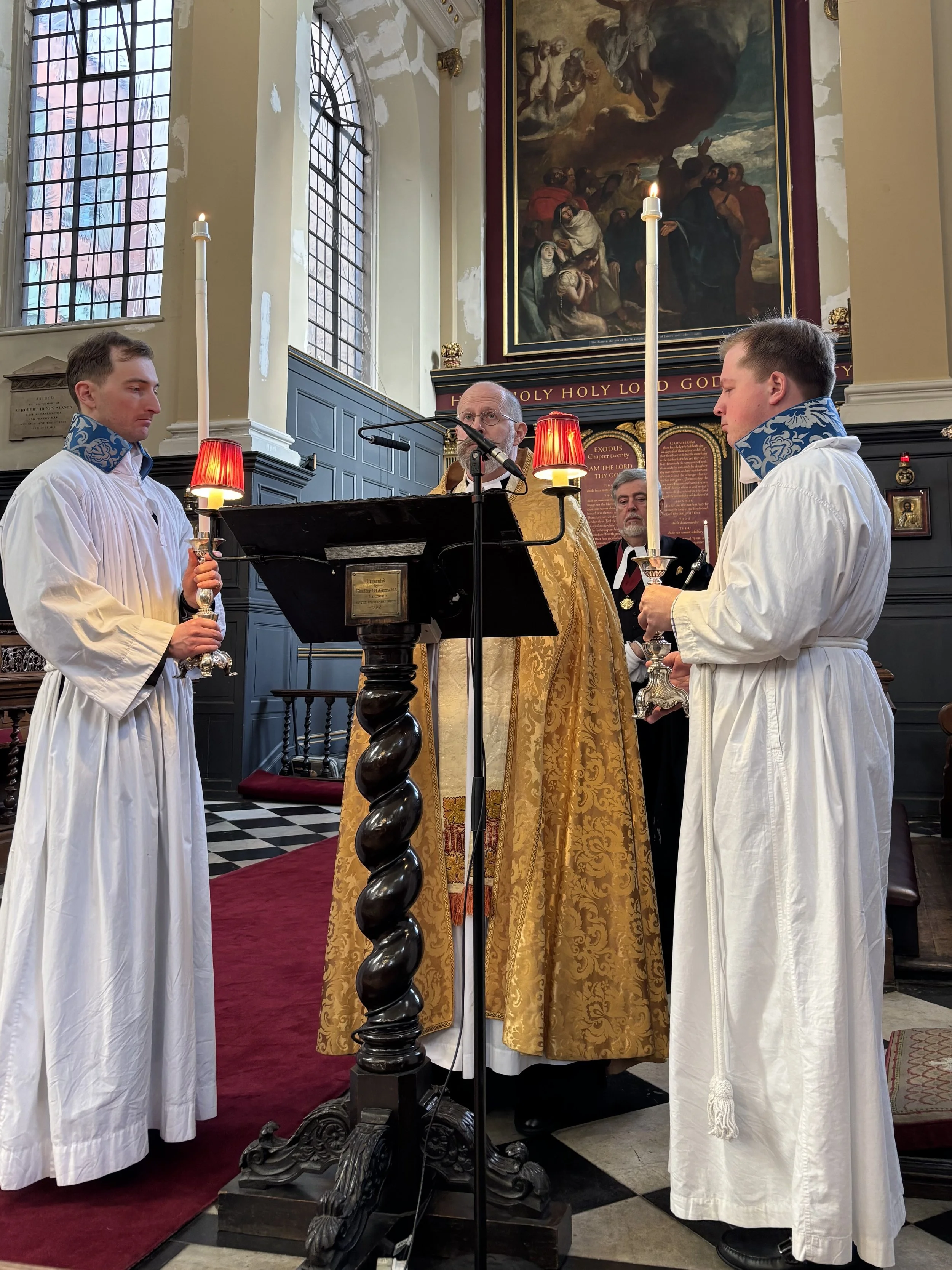 A religious ceremony taking place in a church with two altar servers holding candles, a priest in gold vestments, and a man in the background. The church has tall stained glass windows and religious paintings on the walls.