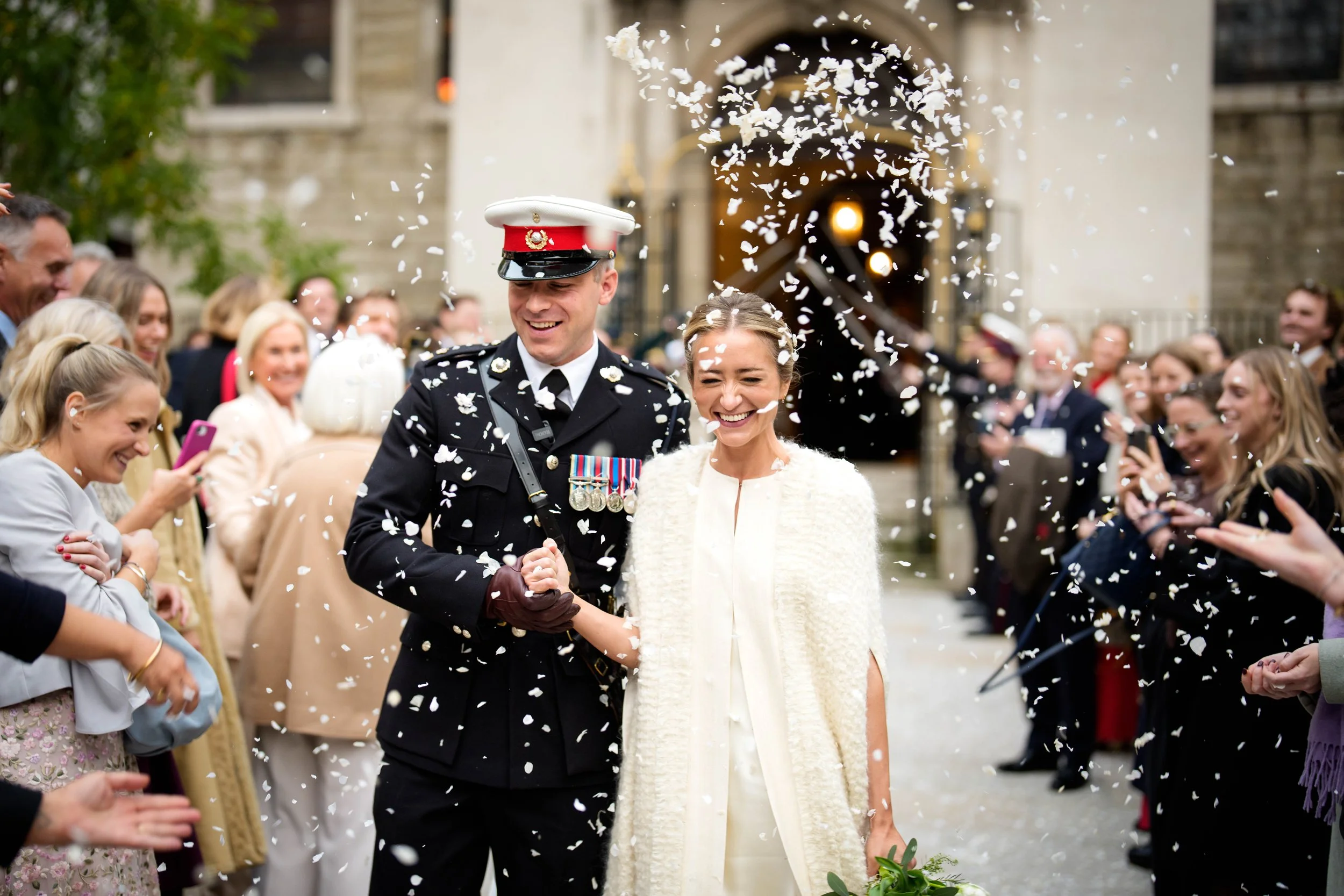 A newlywed couple, with the groom in a military uniform, holding hands and smiling. They are surrounded by guests throwing confetti during a wedding celebration outside in front of a church.