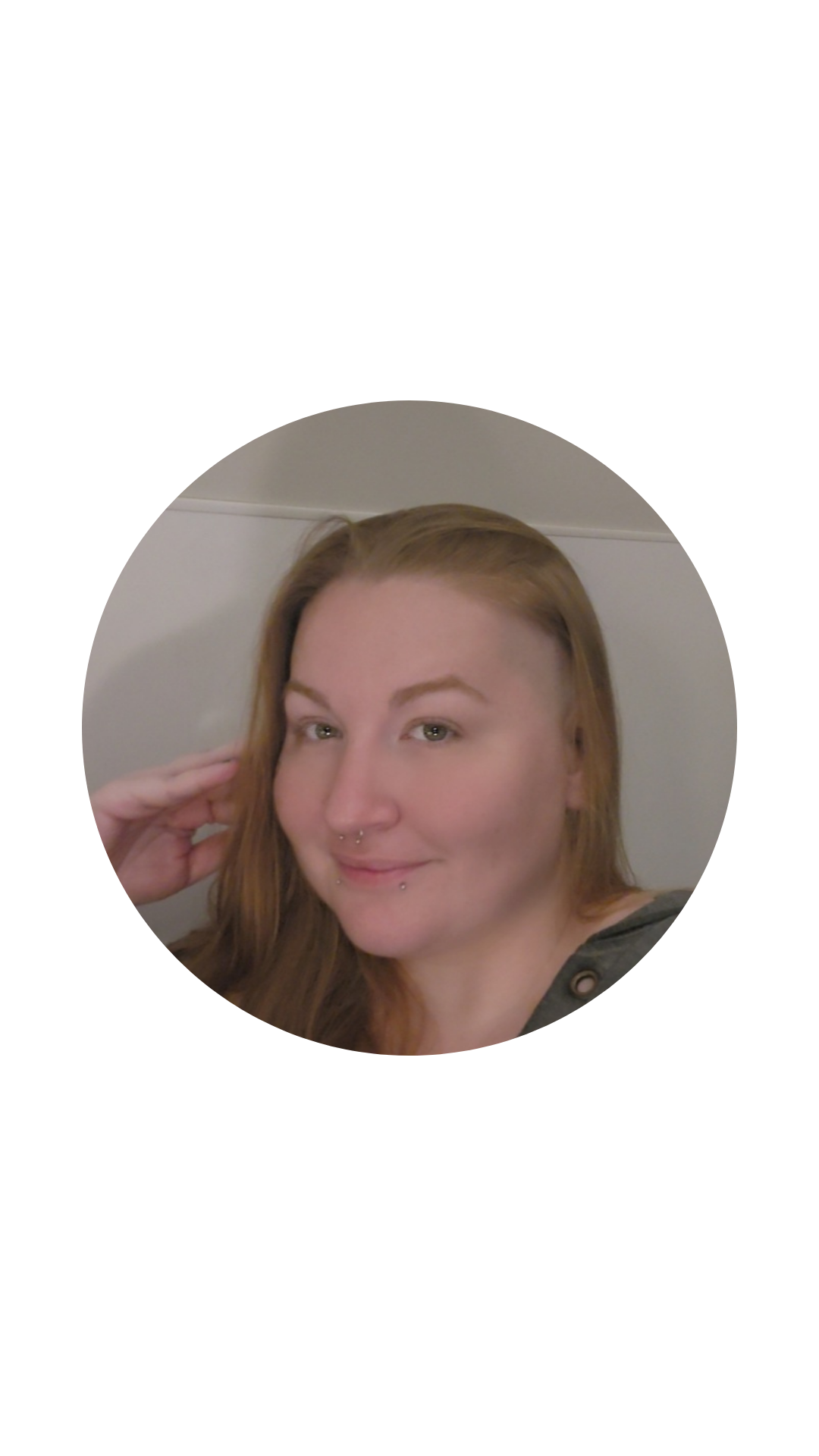 A young woman with light skin, long reddish-brown hair, and multiple facial piercings, including a septum and labret, smiling softly with her hand near her face.