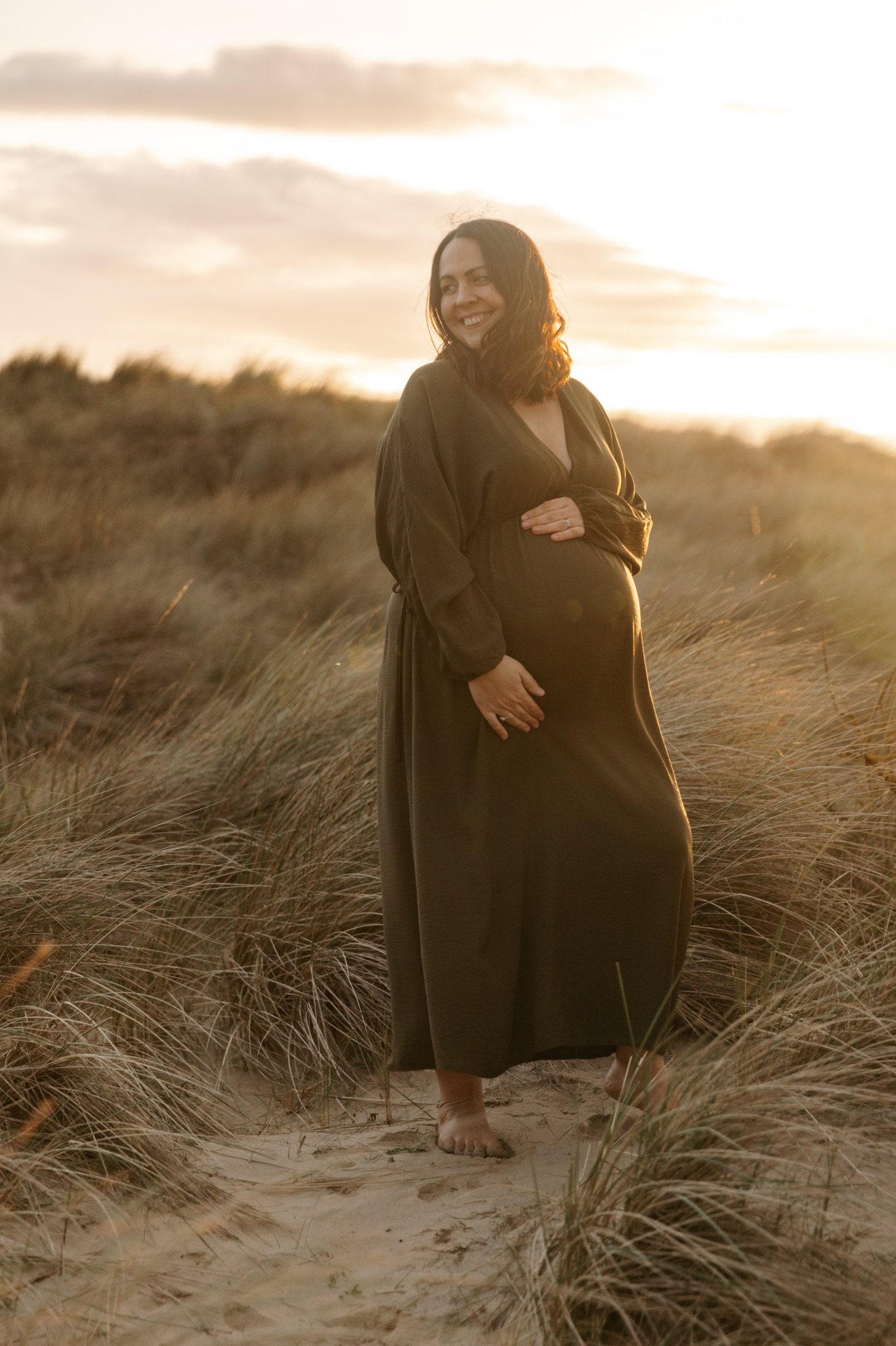 A pregnant woman standing barefoot on the sandy beach, surrounded by tall grass, during sunset, smiling and looking to her left.