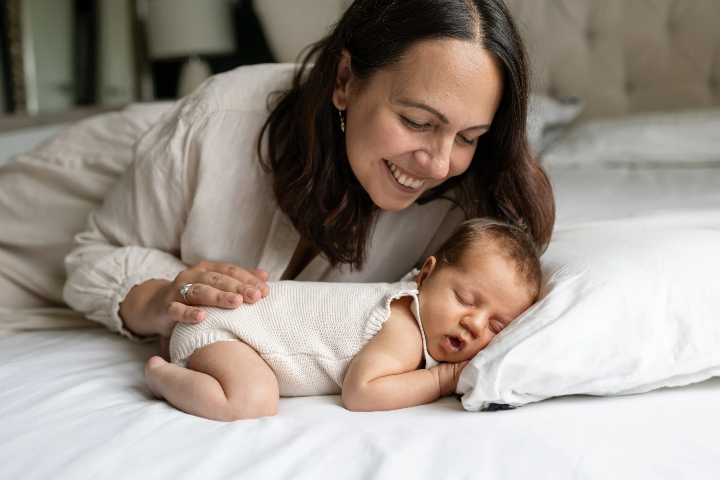 A woman smiling as she leans over a sleeping baby on a bed.