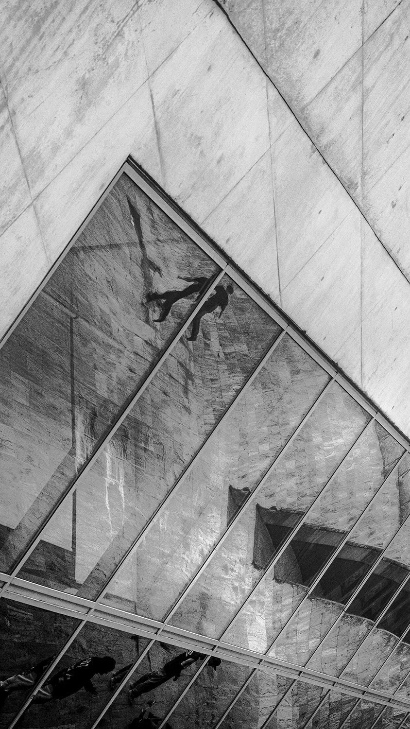 Schwarz-Weiß-Foto einer modernen Treppe mit großen Glasfenstern und reflektierenden Flächen. Mehrere Menschen gehen die Treppe hinauf und hinab.
