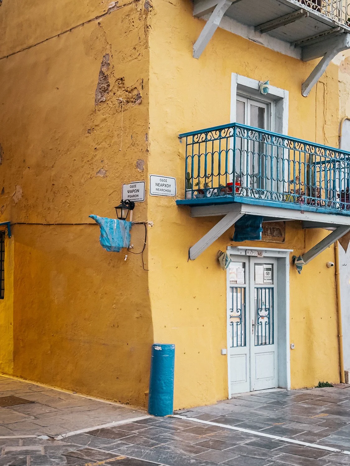 Ein gelbes Gebäude mit blauer Balkonbrüstung und weißer Tür. Zwei Straßenschilder auf der Wand, eine Laterne und eine blauer Pfeiler im Vordergrund