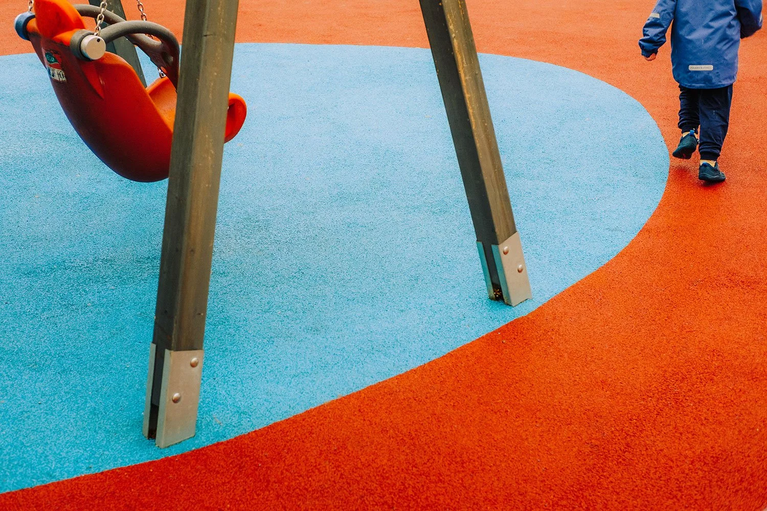 Klettergerüst auf einem Spielplatz mit blauer und roter elastischer Oberfläche, im Hintergrund ein Kind in blauer Jacke, das weggeht.