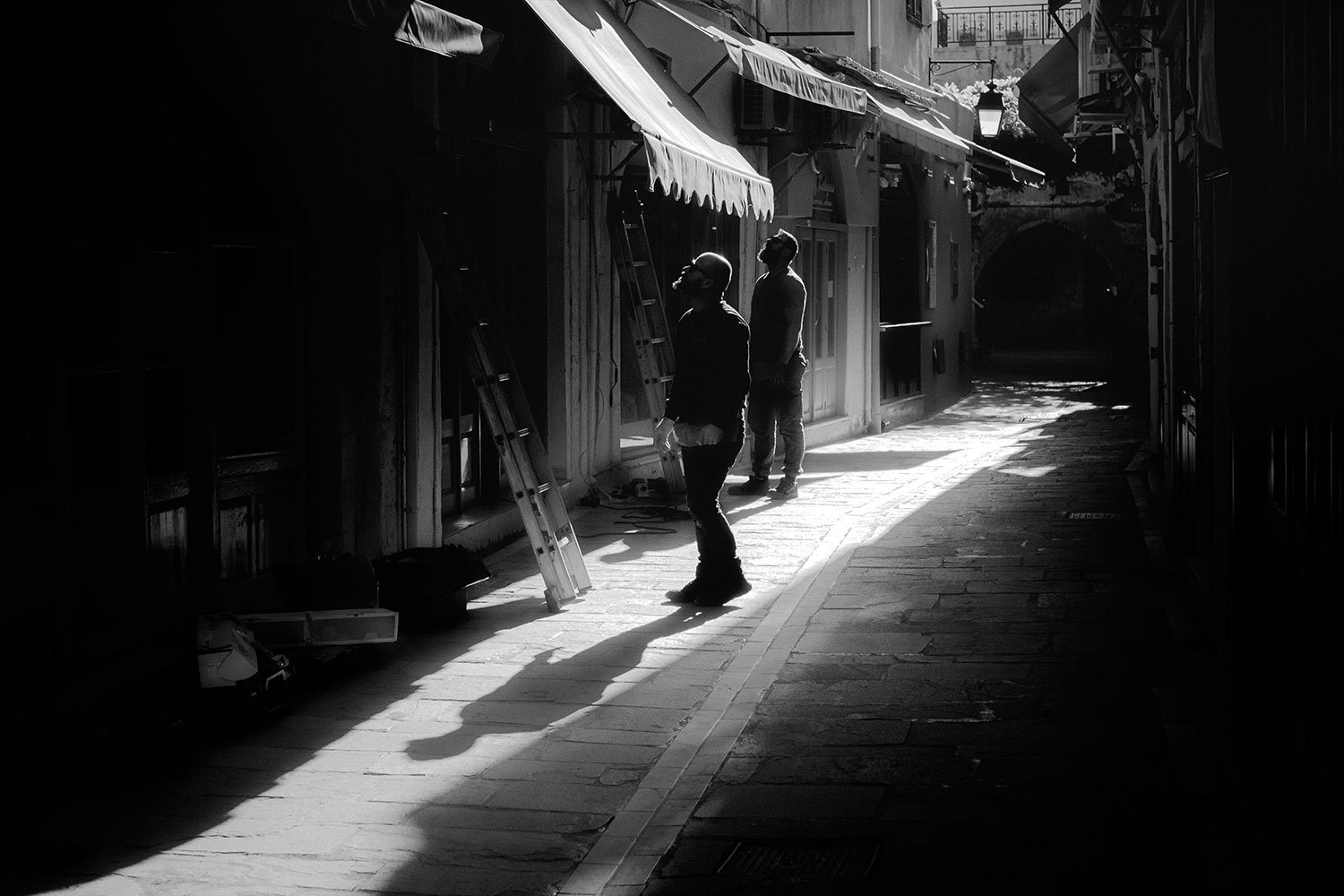 Zwei Menschen stehen in einer schmalen, von der Sonne beleuchteten Gasse, eine Person trägt eine Brille und schaut nach oben, die andere Person steht auf einer Leiter.