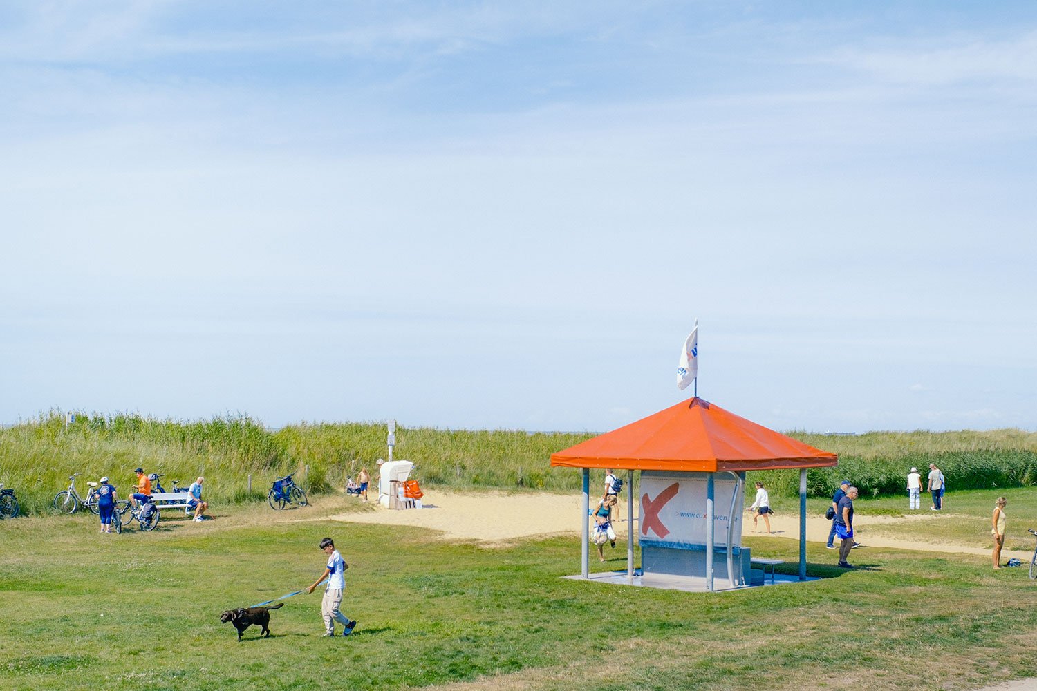 Strandpavillon mit orangem Dach, im Hintergrund Spaziergänger und Fahrradfahrer, Kinder und Hunde auf einer Wiese, Sandstrand und Dünen, blauer Himmel, friedvolle Sommertagesstimmung.