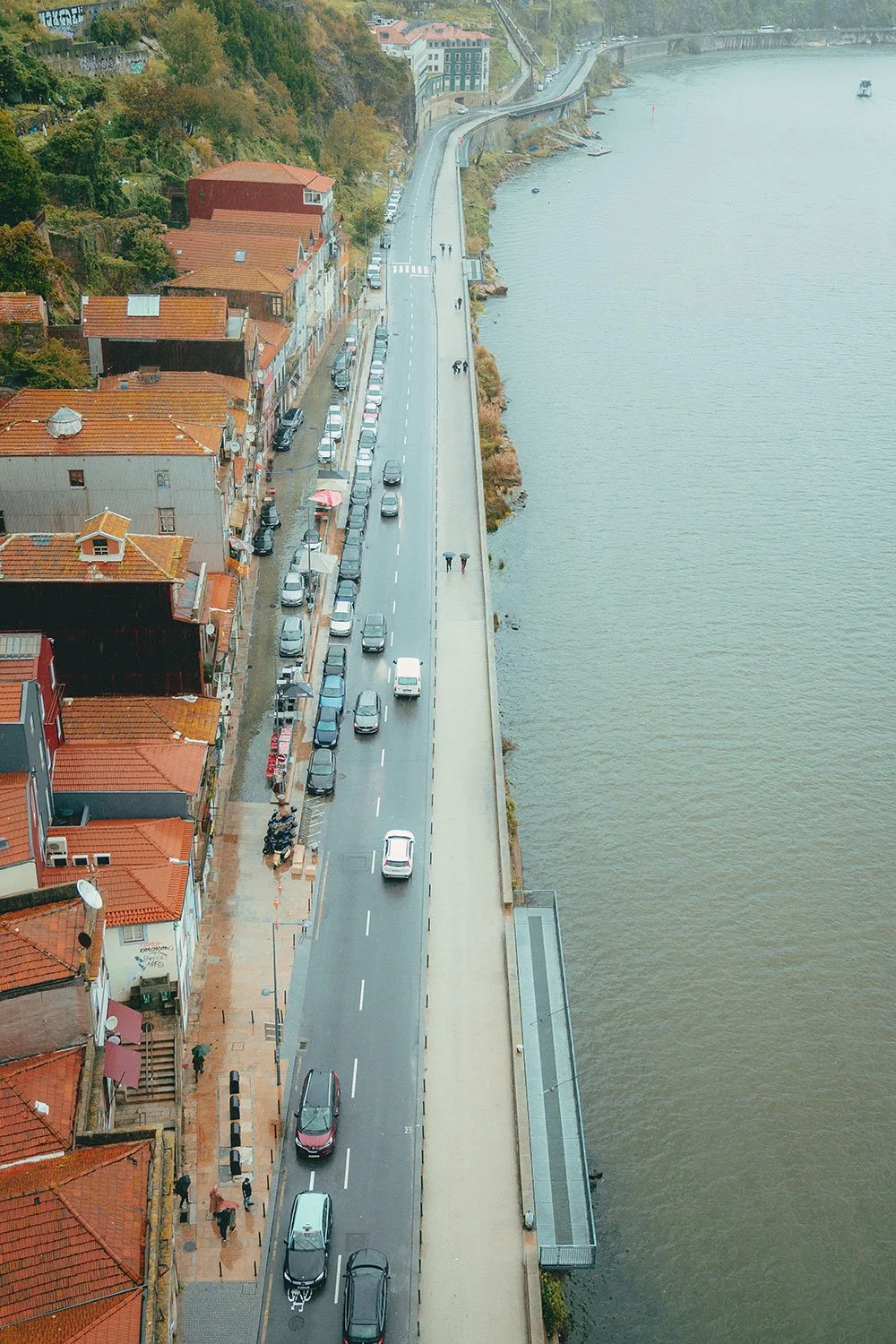 Blick auf eine Küstenstraße mit parkenden Autos, schmalen Gehwegen mit Fußgängern, Häuser mit roten Ziegeldächern auf der linken Seite und einer Gewässerseite mit Wasser auf der rechten Seite, umgeben von Hügeln mit Bäumen.