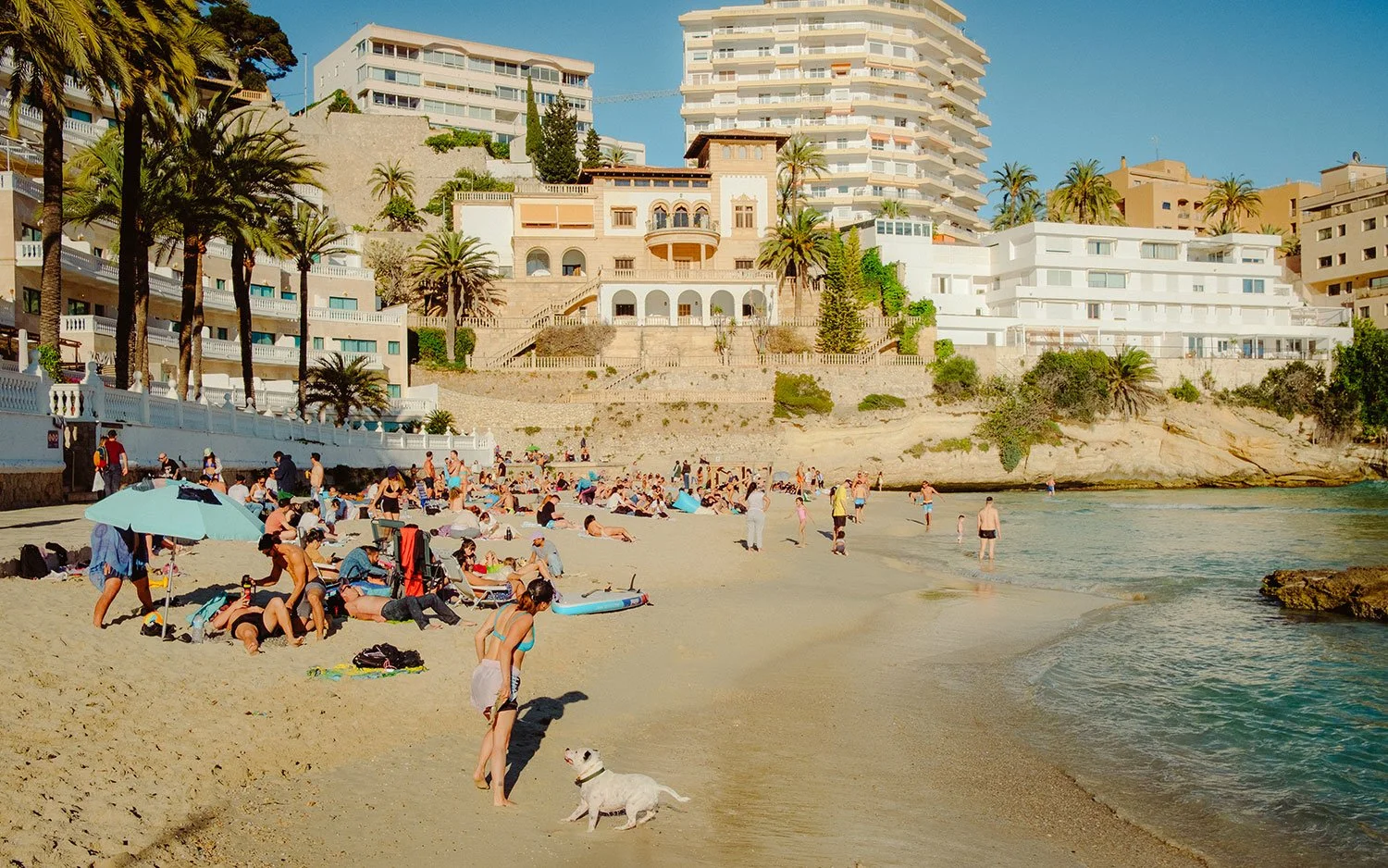 Strand mit zahlreichen Menschen, die sonnenbaden, spazieren gehen und im Wasser spielen, vor einem Hintergrund aus hohen Gebäuden und Palmen.