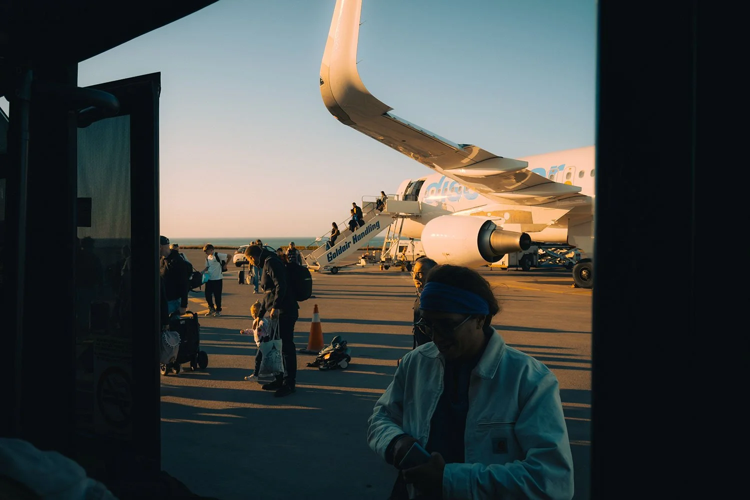 Menschen steigen aus einem Flugzeug aus und gehen auf dem Flughafen. Im Hintergrund ist ein Flugzeug auf dem Rollfeld zu sehen, das bei Sonnenuntergang beleuchtet wird.