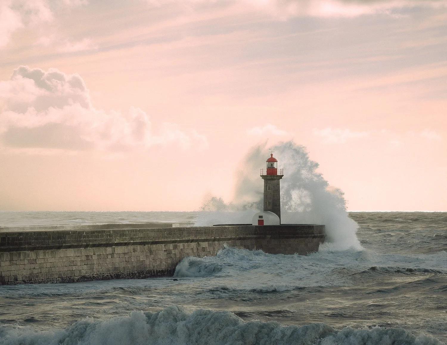 Seehafen bei stürmischem Wetter mit Welle, die gegen den Leuchtturm schlägt, im Abendlicht. Der Leuchtturm ist am Ufer einer Mole. Der Himmel ist leicht bewölkt, in Pastellfarben.